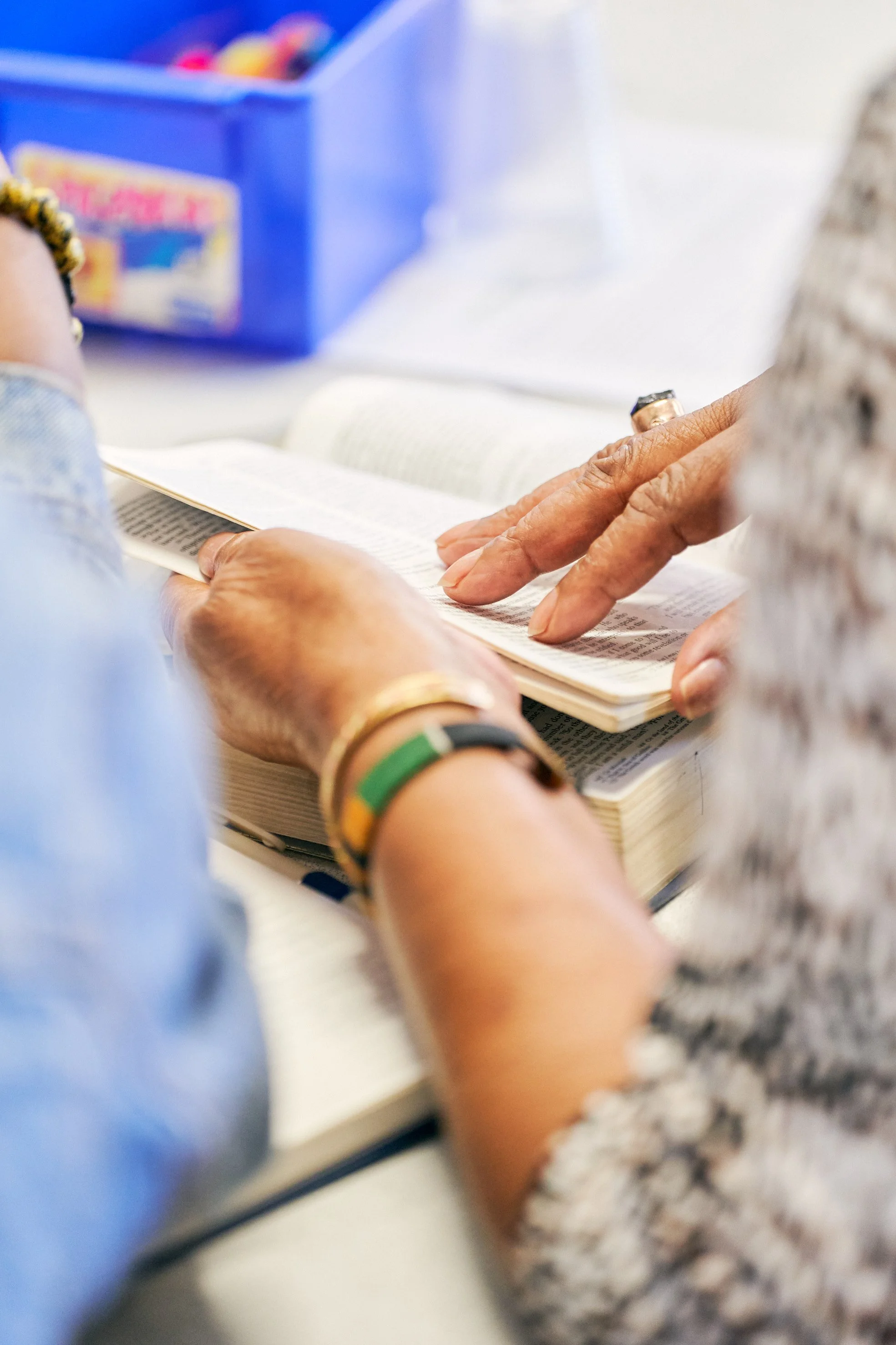 Close-up of hands holding an open book, with a person wearing a bracelet with colored segments and a ring on their finger, and a person wearing a patterned sleeve nearby. In the background, there is a blue box with colorful items inside.