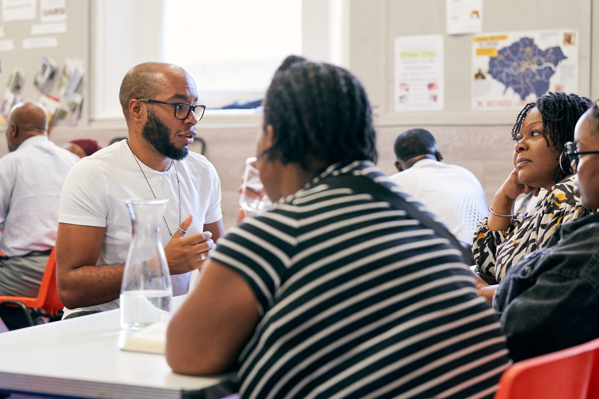 People sitting around a table engaged in a discussion, with a man wearing glasses speaking to a woman in a striped shirt, in a room with a map of Texas on the wall.