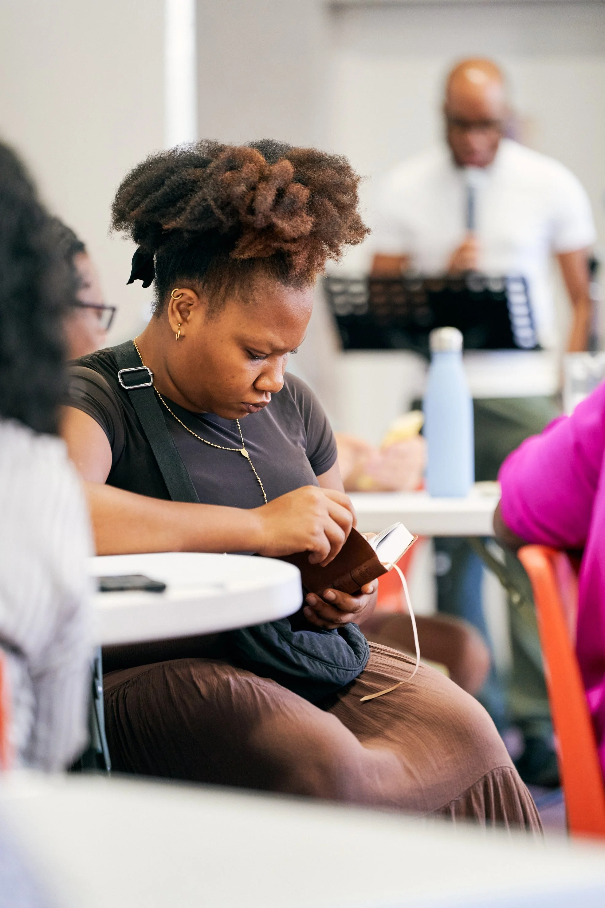 Young woman with natural curly hair reading a book during a meeting; blurred man in the background holding a microphone.