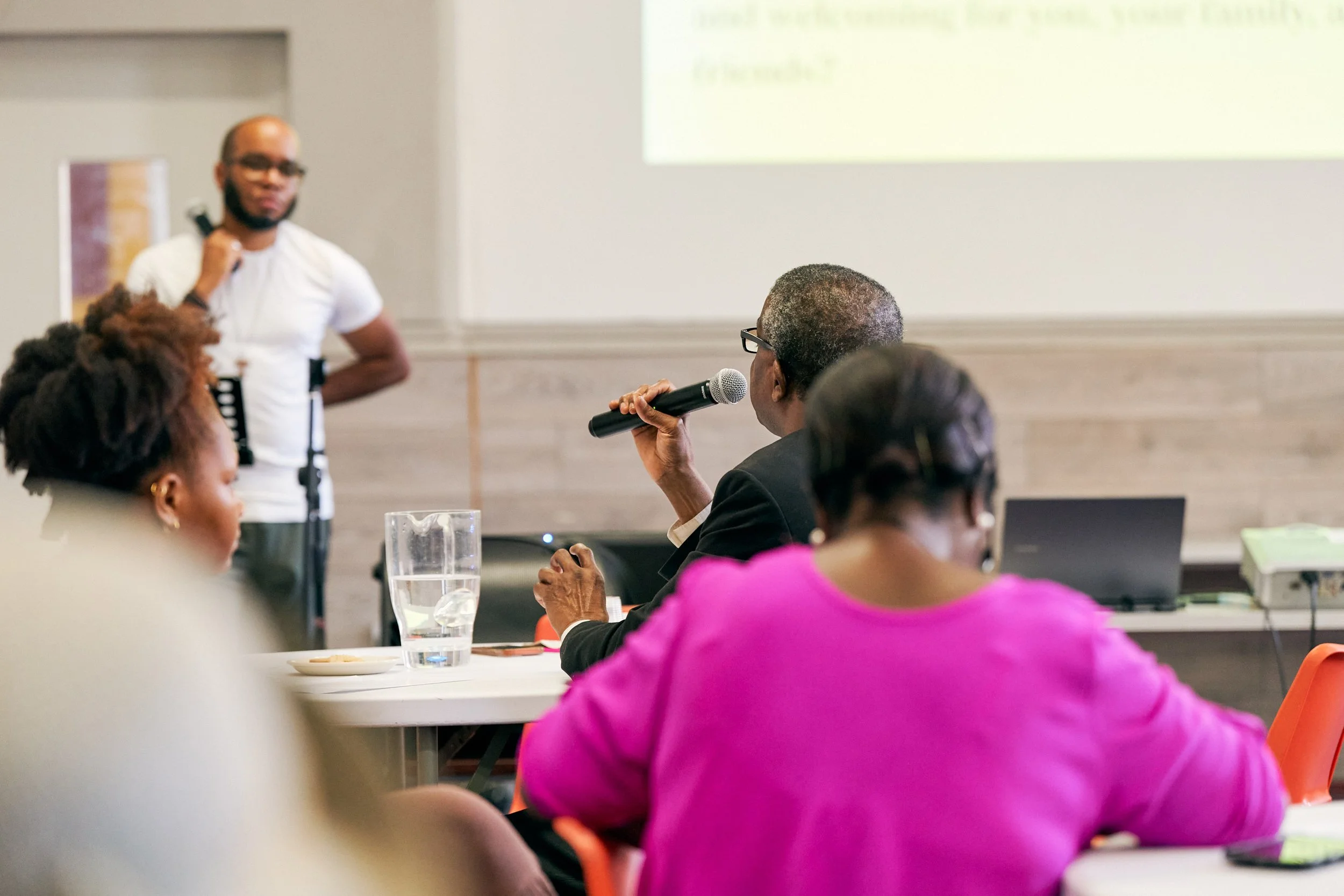 A man with glasses and a beard is standing at the front of a room, holding a microphone in his left hand and a small object in his right hand. Several people are seated at tables, listening, with one woman in a bright pink top in the foreground. Ther