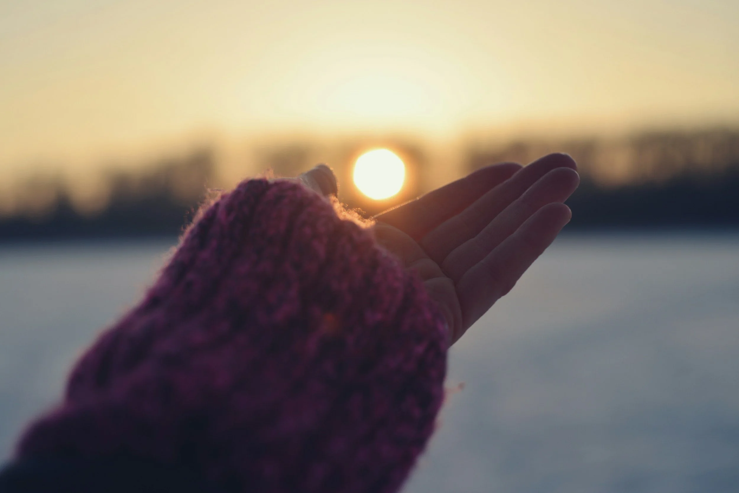 Close-up of a hand with a pink glove reaching towards the setting sun over a body of water at dusk.