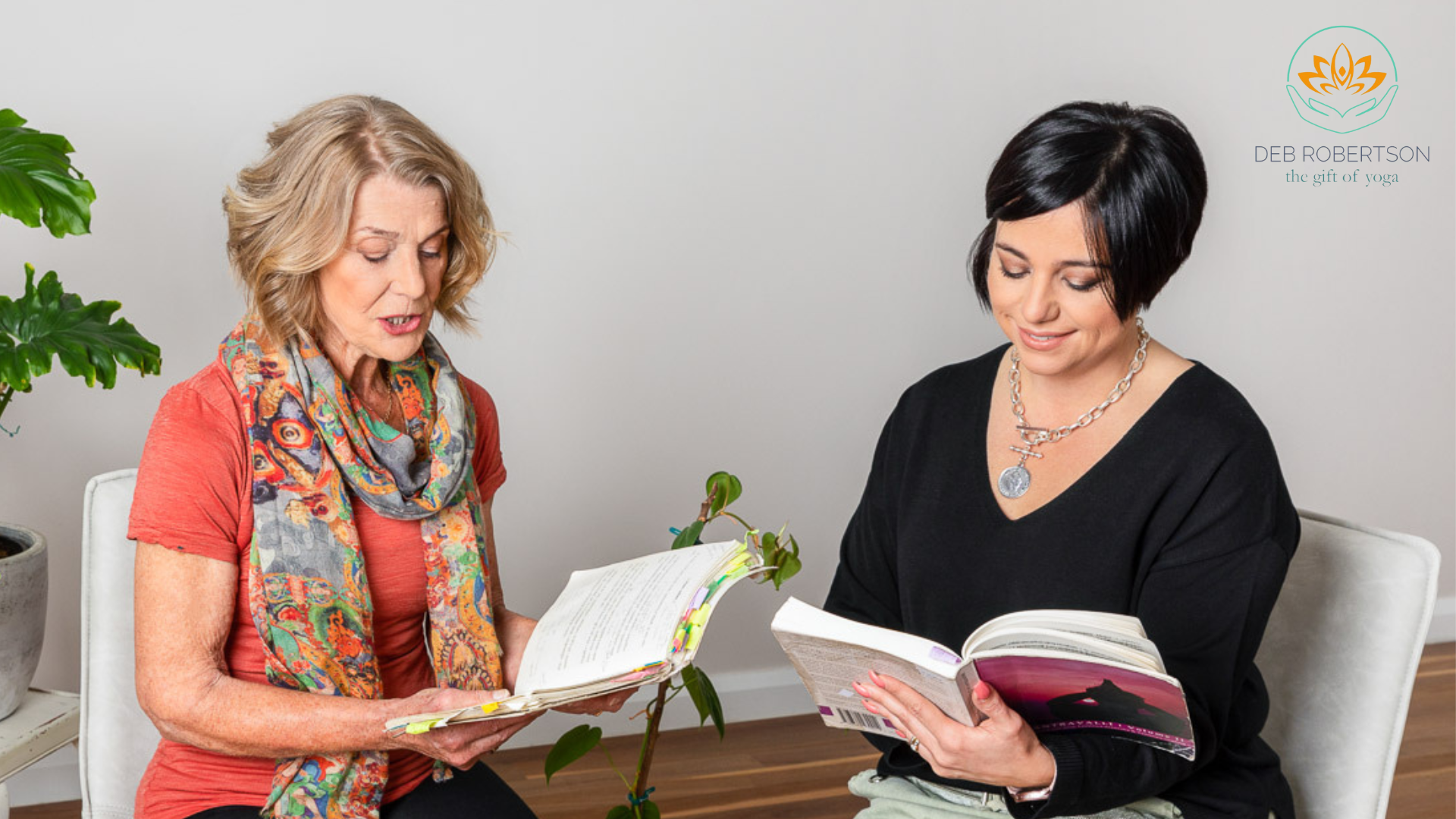 Two women sitting in a room, reading books, with a potted plant nearby and a logo with a lotus and text in the top right corner. Deb Robertson Vedic Chanting