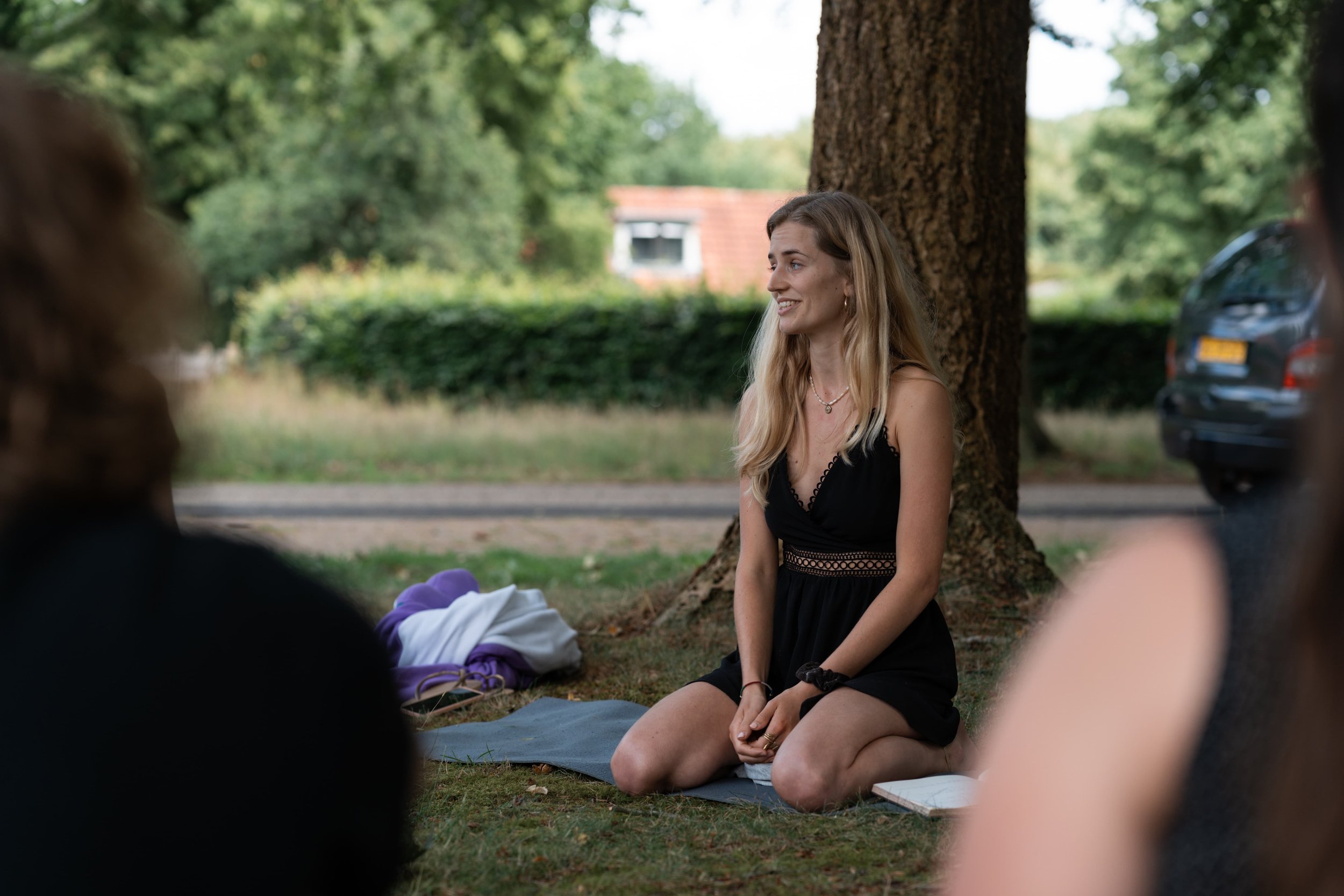 woman sitting on a mat in nature surrounded by people in a black jumpsuit