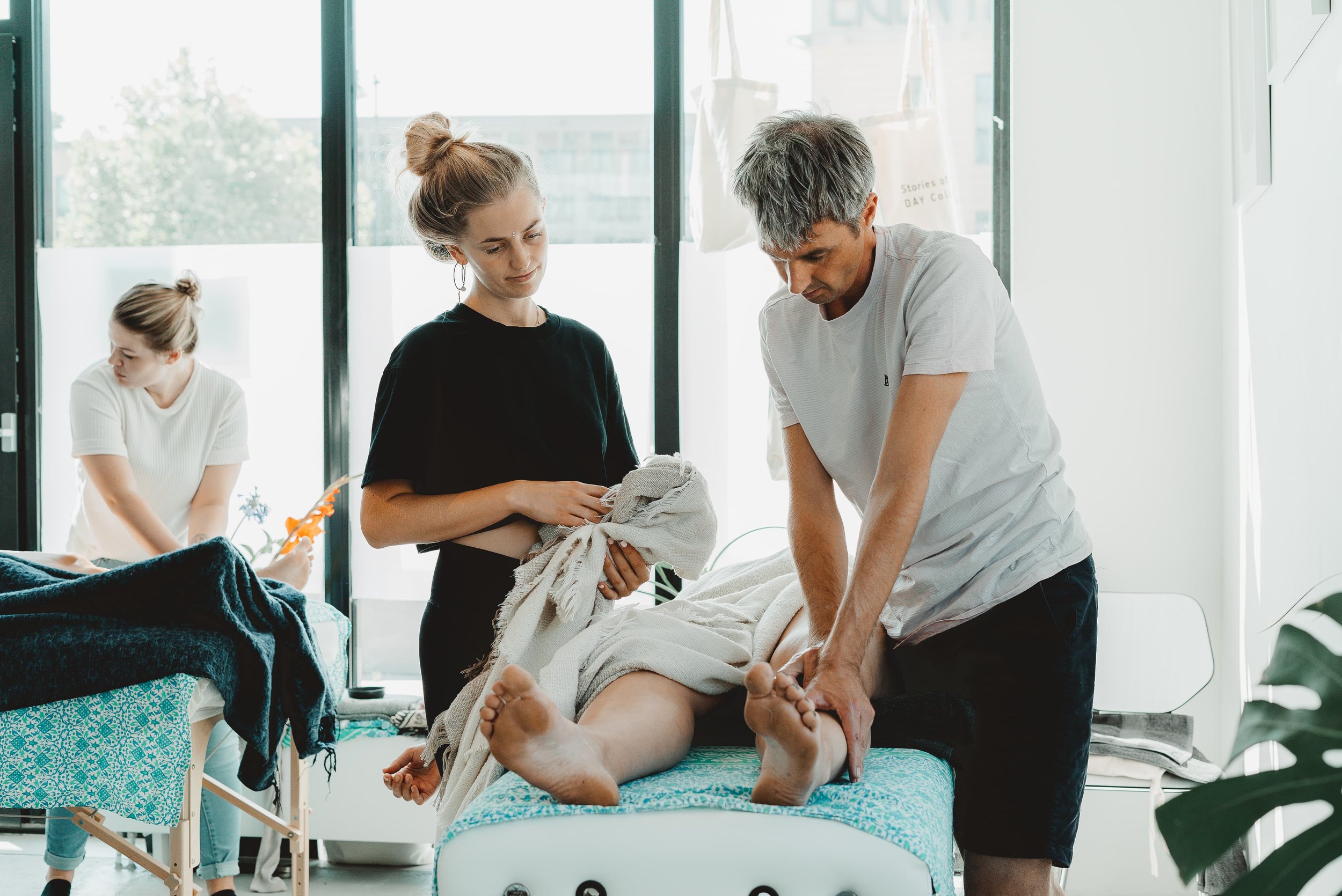 a female massage teacher looking at a male student massaging the legs of a woman