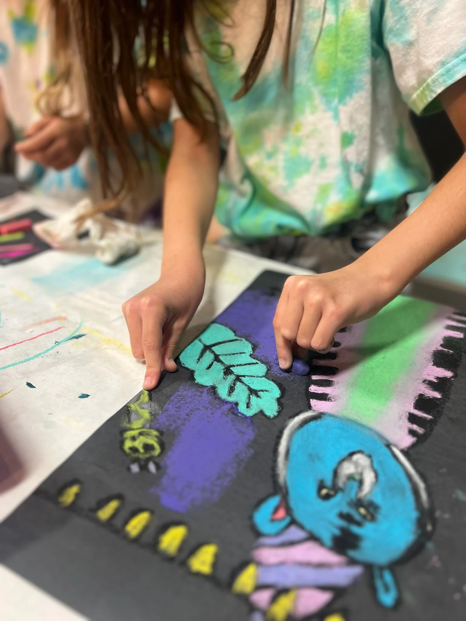 Children creating a colorful chalk drawing on black paper, featuring a blue creature with an eye and a leafy branch.