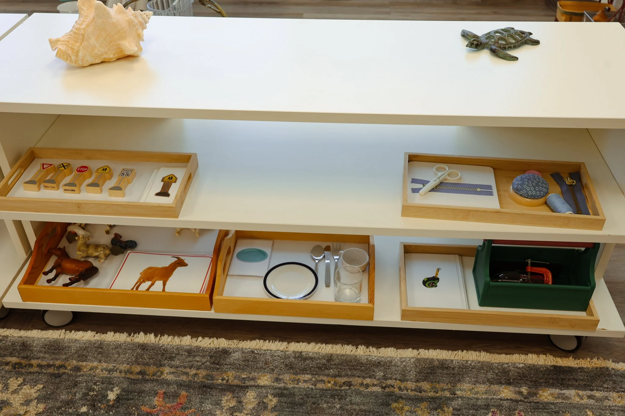 White multi-tiered shelf with wooden trays containing toy animals, miniature furniture, and accessories, arranged on a rug in a room. AMI Montessori Training Center, the Montessori Institute of North Texas