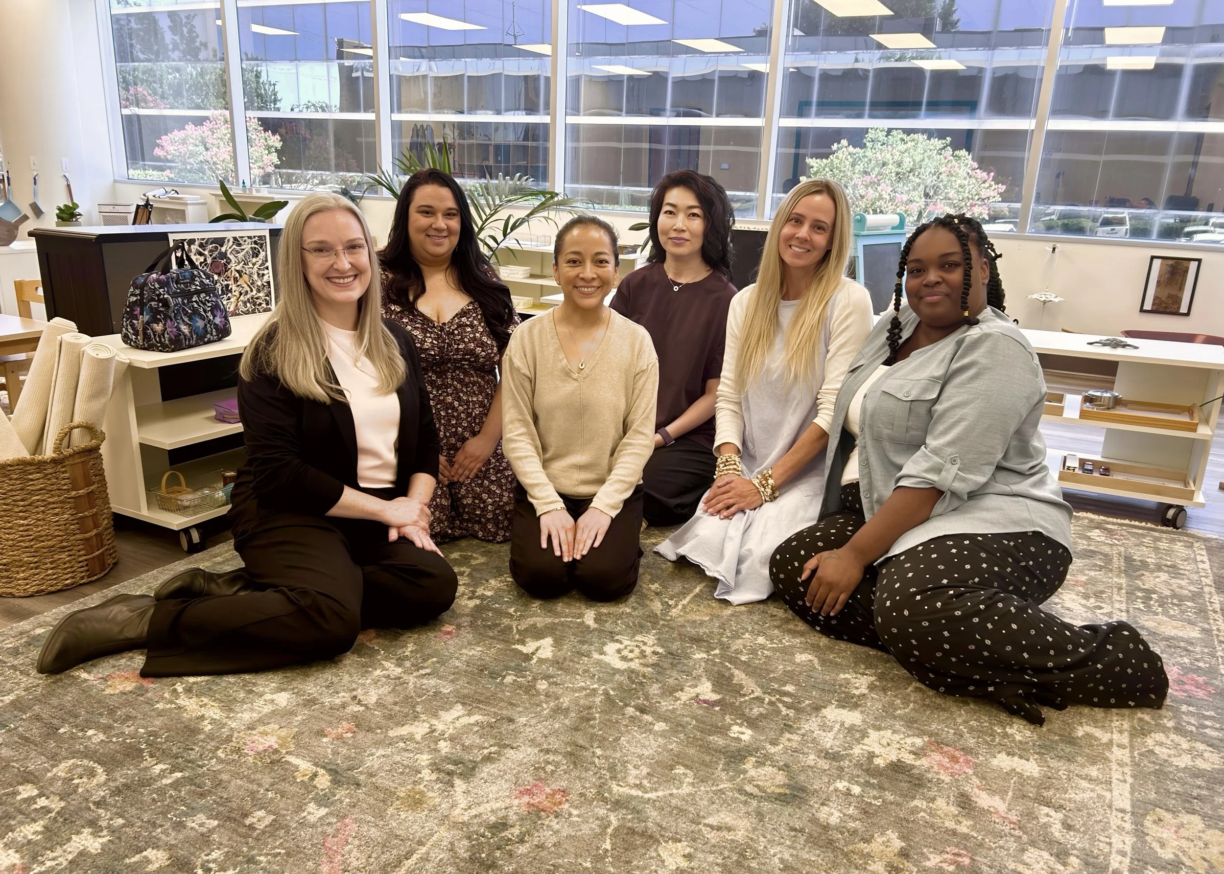Six women sitting and kneeling on a patterned rug in a room with large windows and various items on shelves behind them. AMI Montessori Training Center, the Montessori Institute of North Texas