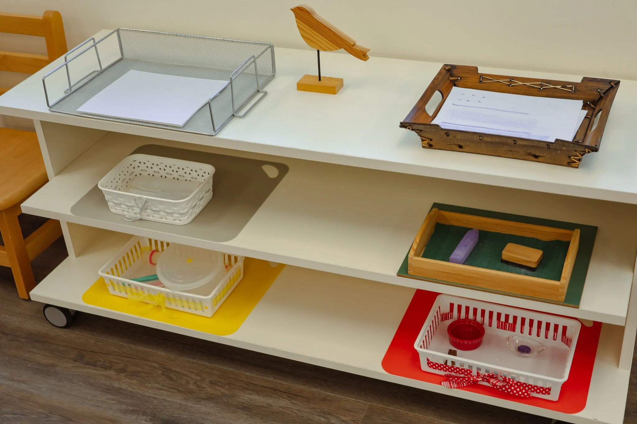 A white shelving unit holding various objects, including empty wire and wooden trays, a wooden lamp, and small containers on colored mats. AMI Montessori Training Center, the Montessori Institute of North Texas