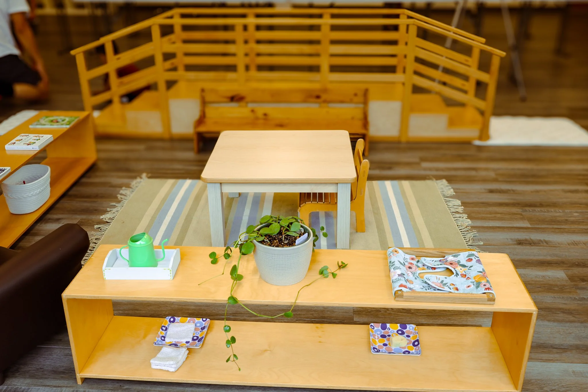 Children's play area with small wooden table, yellow chairs, a potted plant, shelves with books, and a wooden fence in the background. AMI Montessori Training Center, the Montessori Institute of North Texas