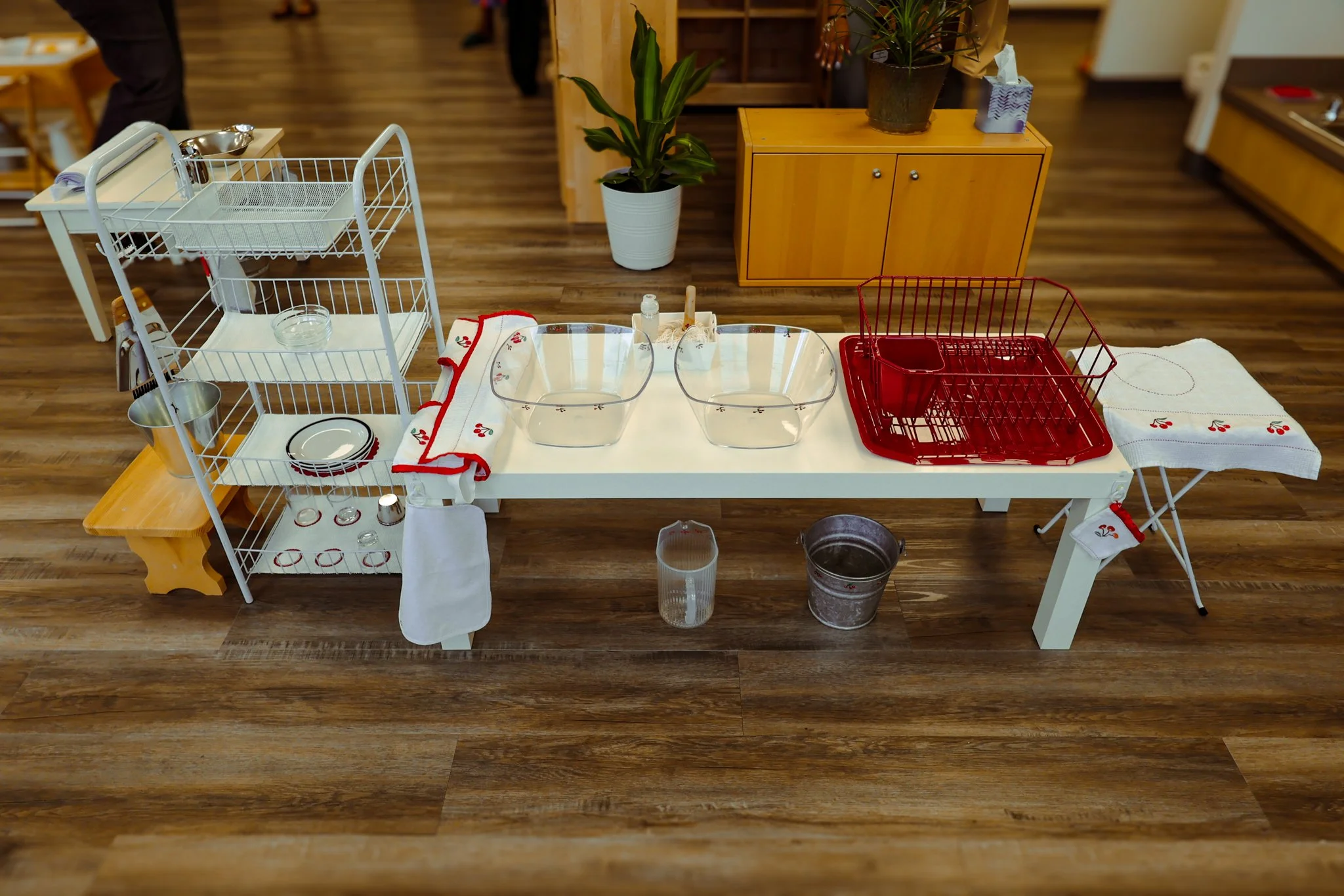 A stack of dish racks with dishes, cups, and utensils, a large glass bowl, a red dish drying rack, and cloth towels on a white table in a room with wooden floors and a small wooden cabinet. AMI Montessori Training Center, the Montessori Institute of