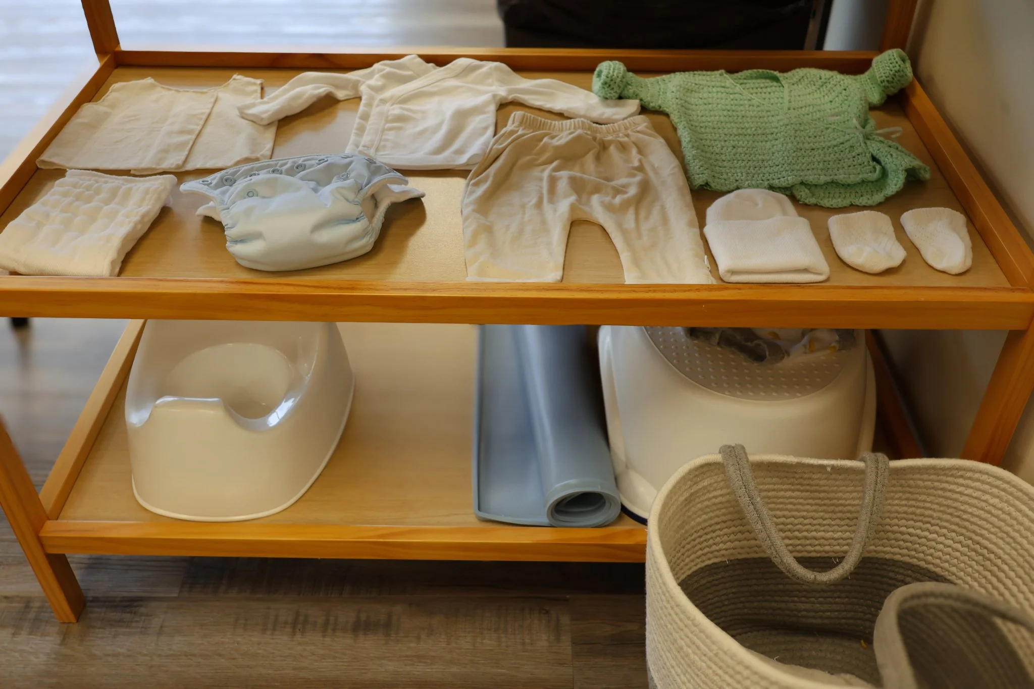 Change table with baby clothes, socks, and a green knitted sweater on top. Below are a baby high chair, a gray roll of paper, and a woven storage basket. AMI Montessori Training Center, the Montessori Institute of North Texas