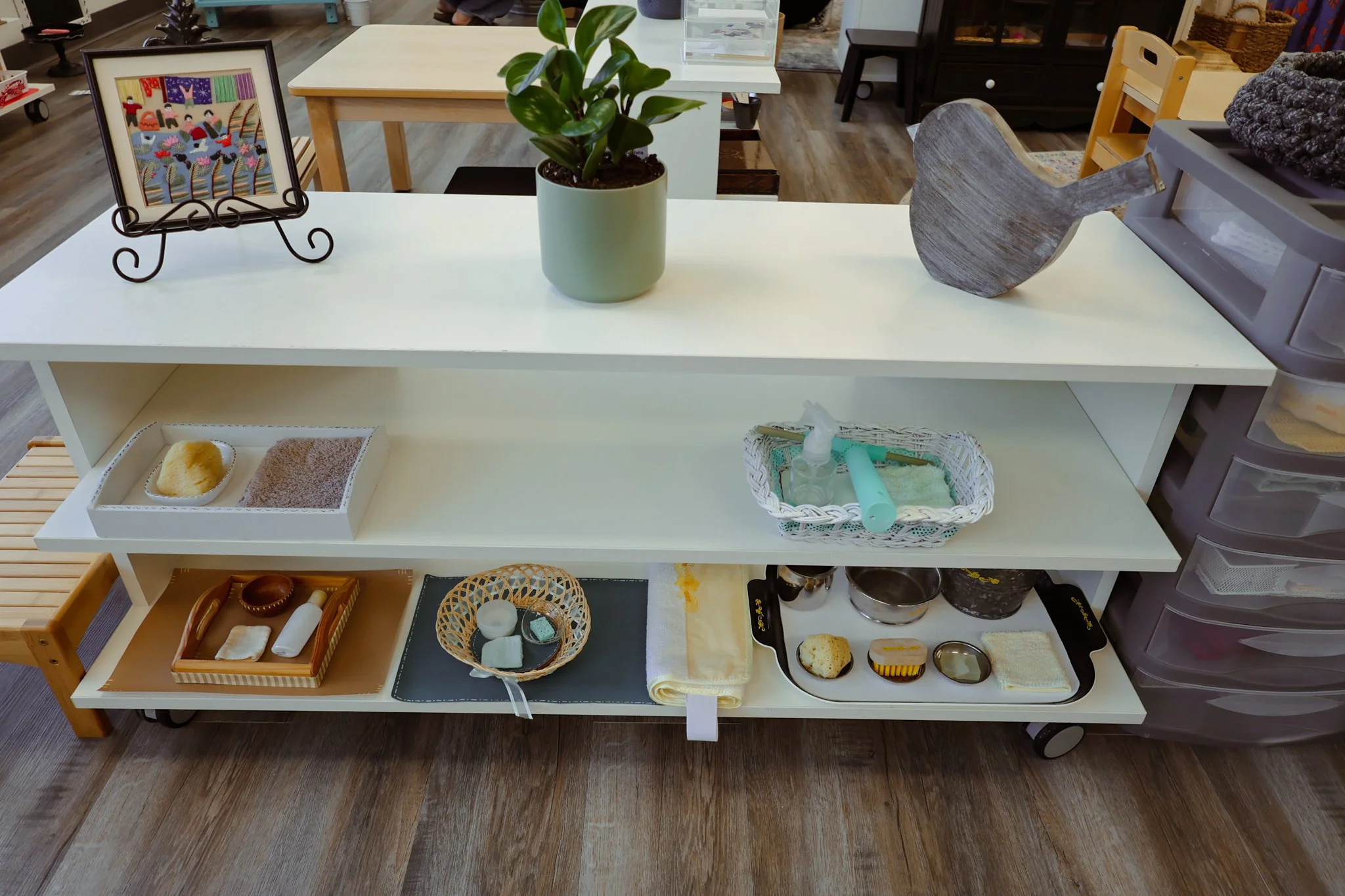 A white multi-level storage cart with various items, including a green potted plant, a wooden whale sculpture, and a framed embroidered artwork on a stand. Shelves hold bathroom items, a towel, soap, and small containers. In the background, there are