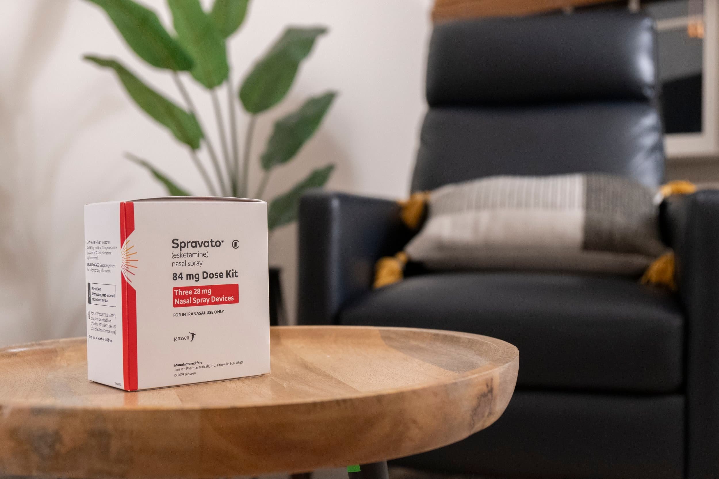 A Spravato nasal spray kit for treatment resistant depression rests on a wooden table near a black armchair and green potted plant.
