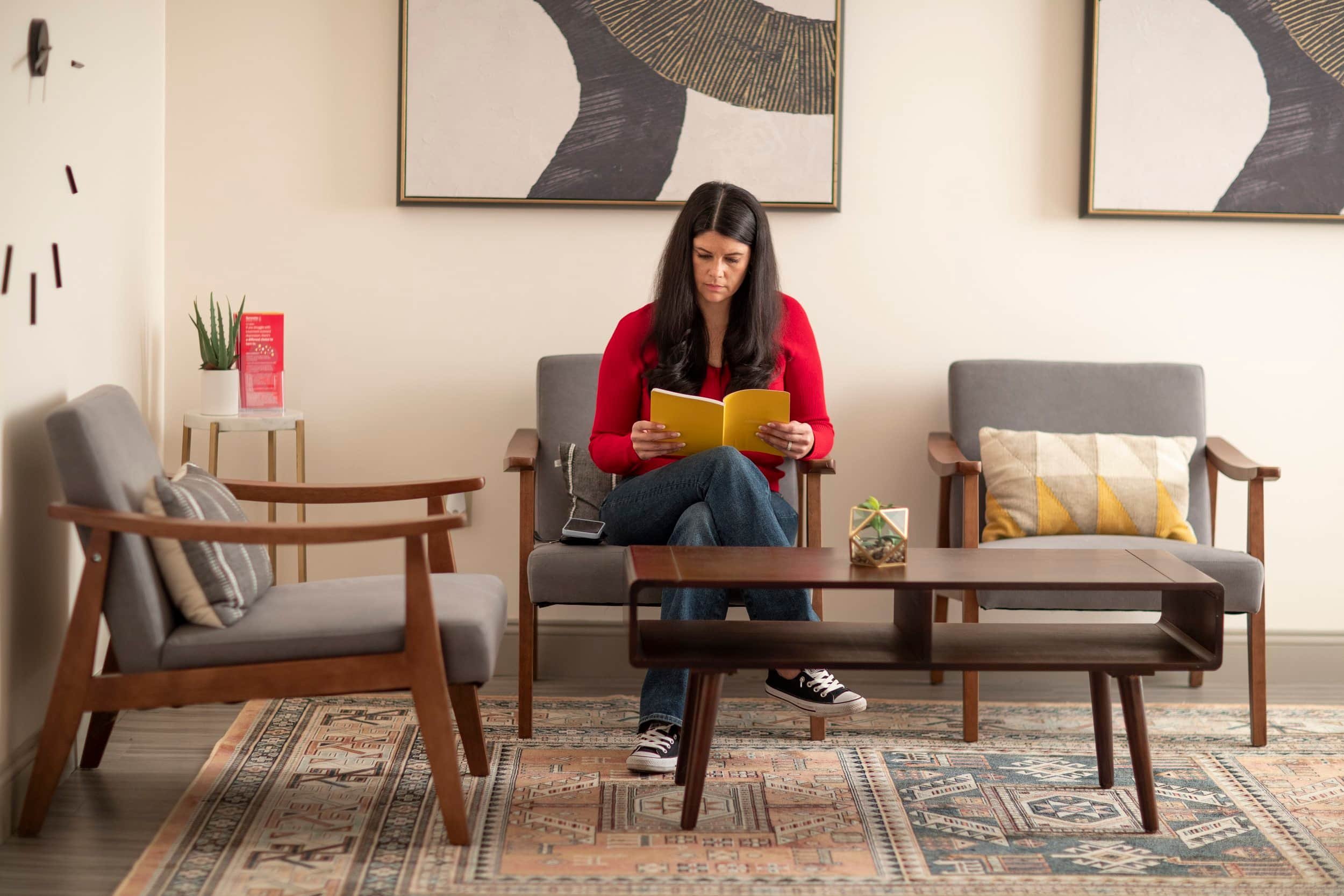 Woman in a red sweater sits on a gray chair reading a yellow book in a modern, cozy living room with mid-century decor.