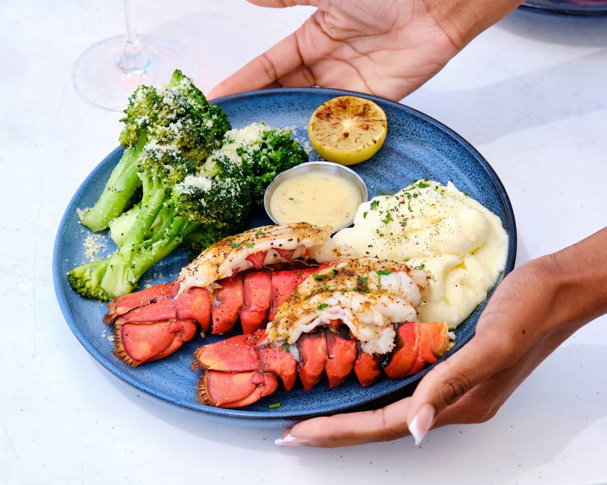 Hands holding a plate with lobster tails and sides at Ocean & Acre