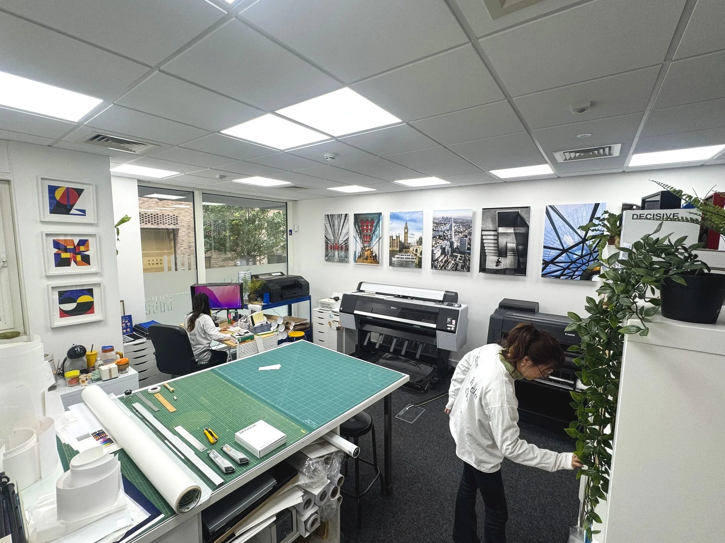 An office with two women working; one is sitting at a desk with a computer, and the other is standing by a white cupboard with a leafy plant on top. The office is decorated with framed abstract art and cityscape photographs on the walls, and contains printing equipment and a cutting table.