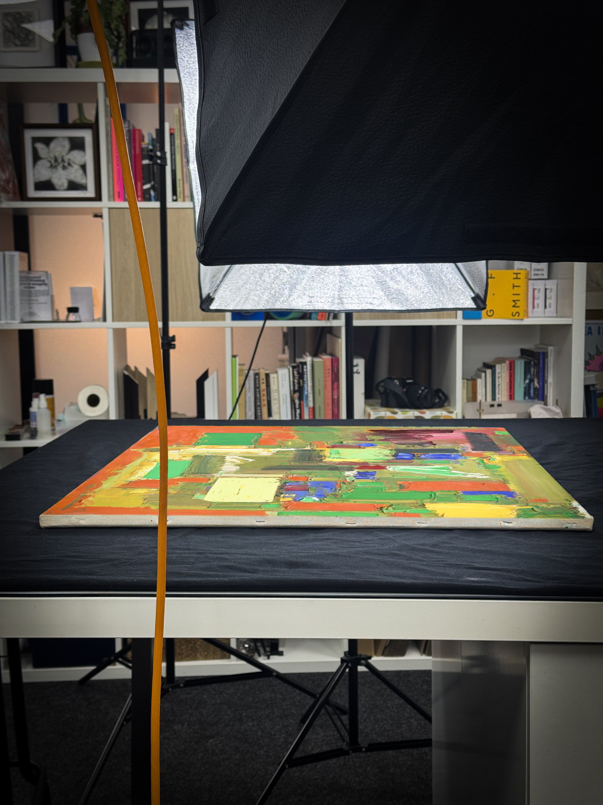 An abstract colorful painting on a canvas placed on a table with photography equipment illuminated by a softbox light in a room with bookshelves.