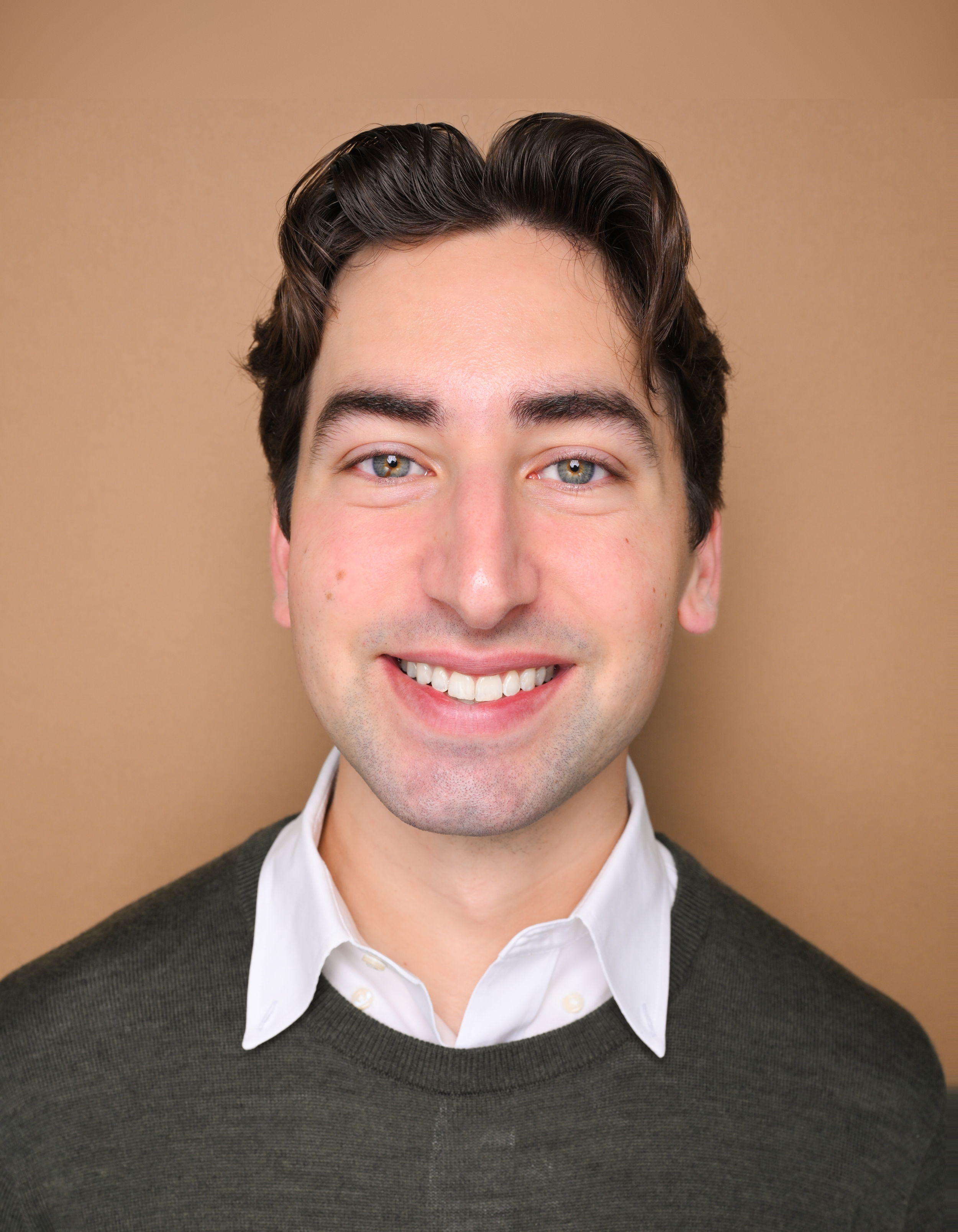 Close-up of a young man with brown hair and blue eyes smiling, wearing a white collared shirt and dark sweater, against a beige background.