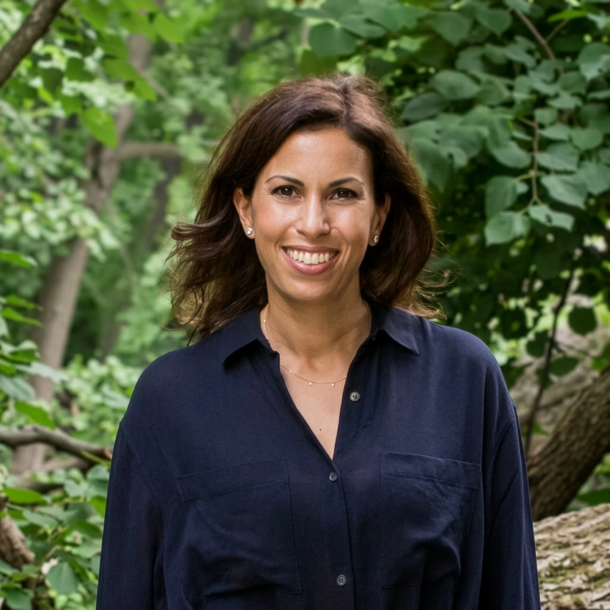 Close-up of a woman smiling outdoors, wearing a navy blue shirt and earrings, with lush green leaves in the background.