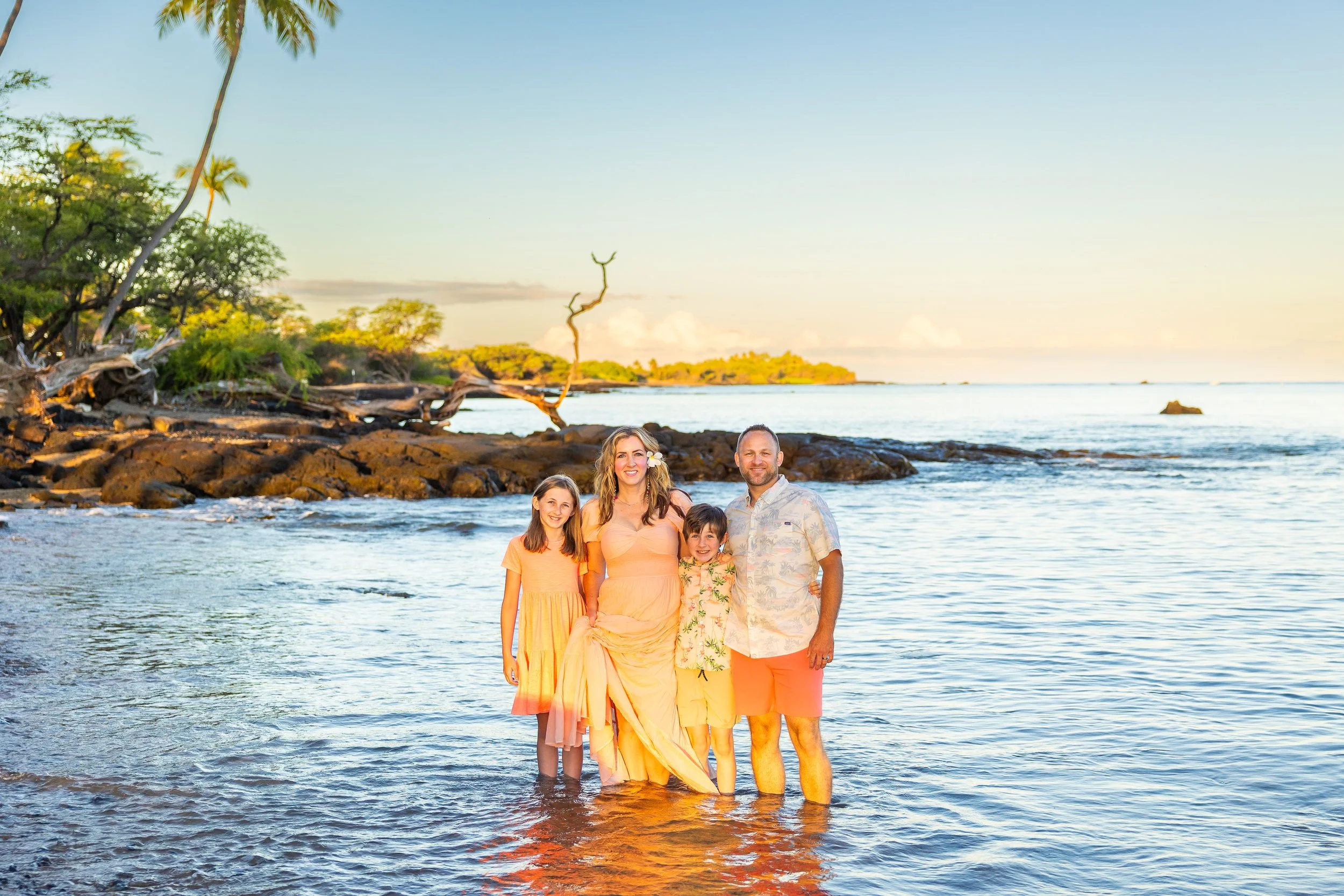 Family standing in the ocean for a sunrise family photoshoot. There are lava rocks and palm trees in the background with the sunrise in the far background.
