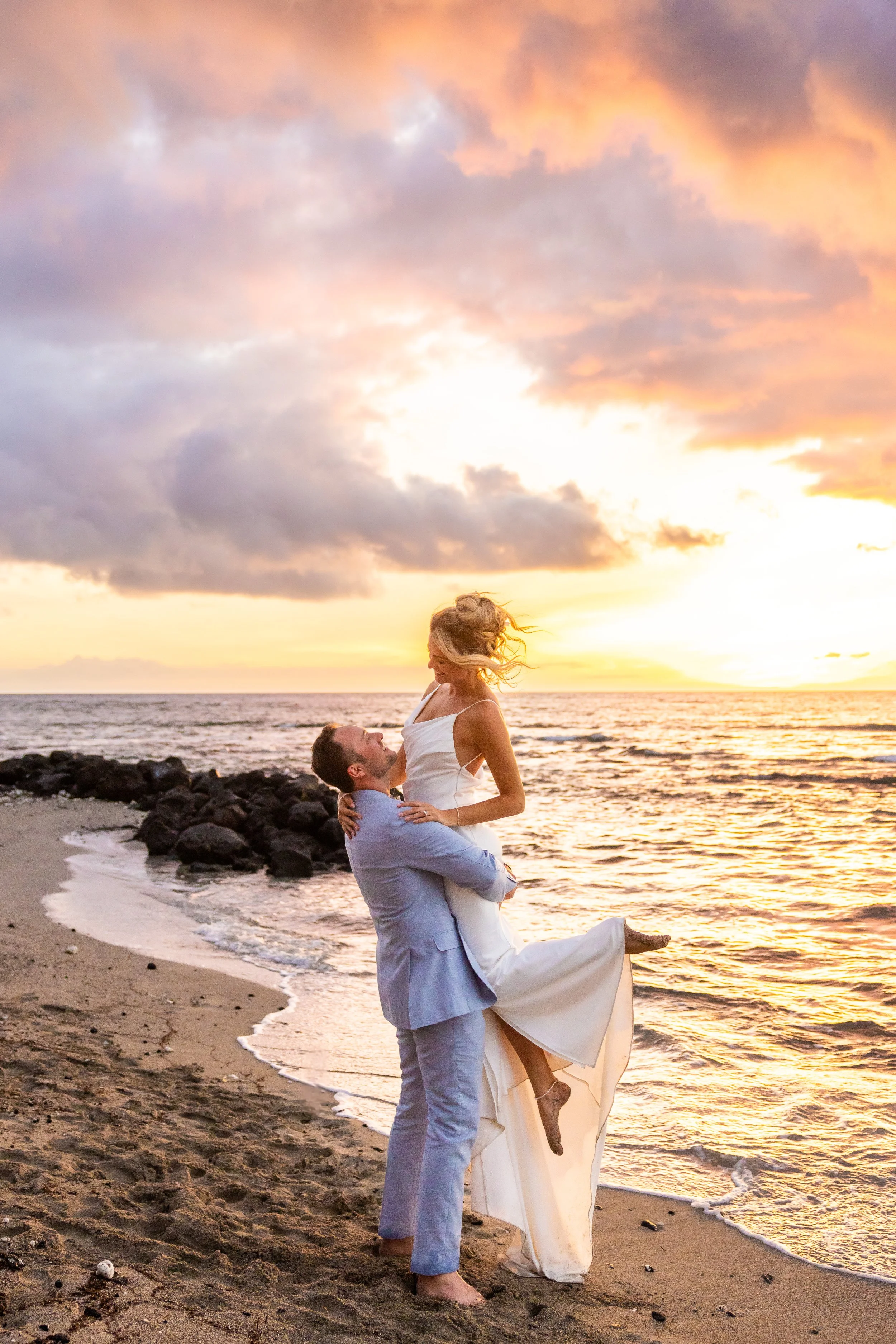 A groom in a light blue suit lifts a bride wearing a long white dress into the air while spinning her. They are on a white sand beach with lava rock on the coast of Hawaii Island. A vibrant sunset is in the background.
