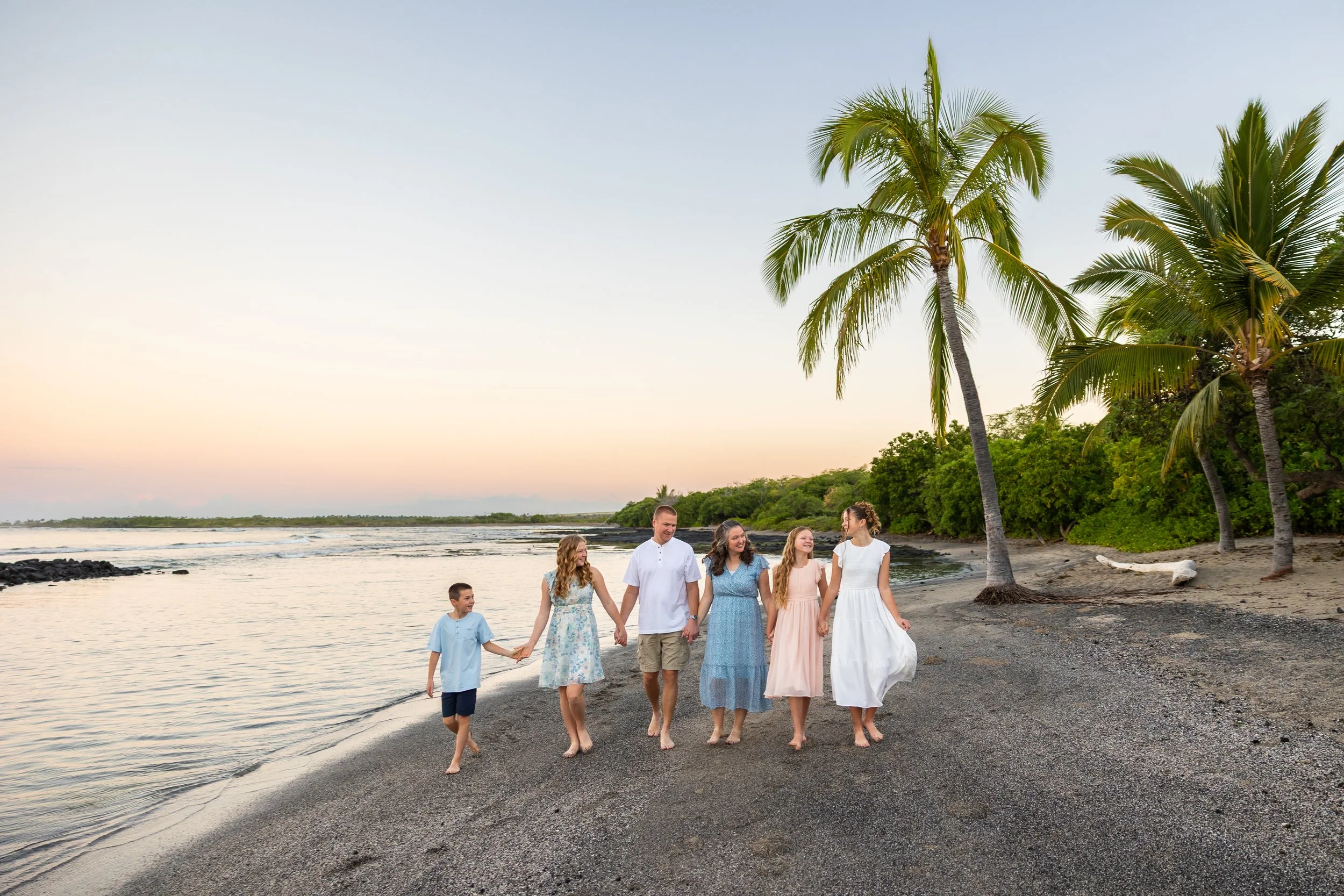 Family getting their photos taken walking towards the camera on a black sand beach with palm trees and the sunrise in the background.