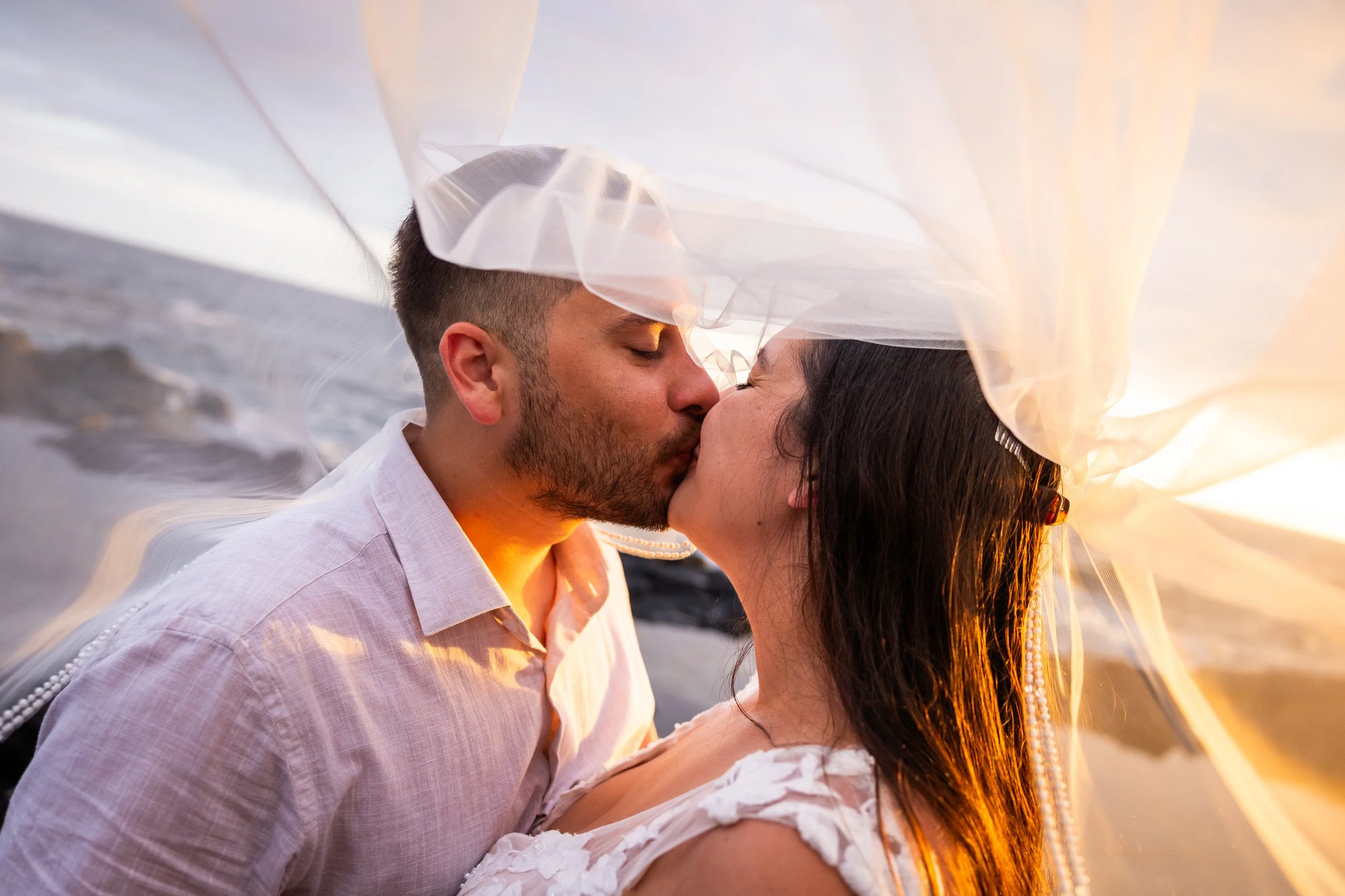 A couple sharing a kiss under a flowing piece of fabric during sunset.
