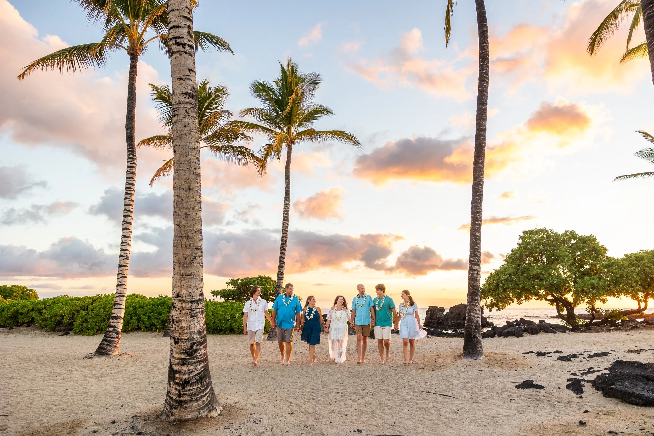 A family getting their photos taken at a beach on the Big Island of Hawaii. The family is walking towards the camera while the sun is setting in the background. They are walking on white sand with green shrubbery and palm trees are in the background.
