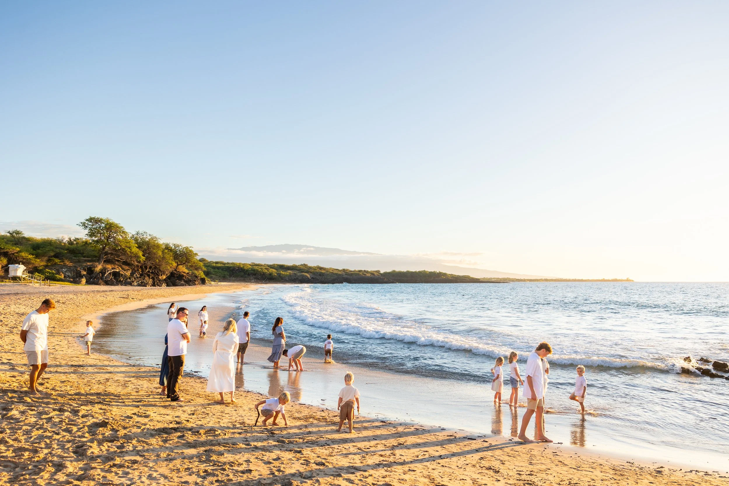 A family playing together on a long white sand beach. They are looking out over the ocean at sunset. Palm trees are in the background as well as the mountain Hualalai.