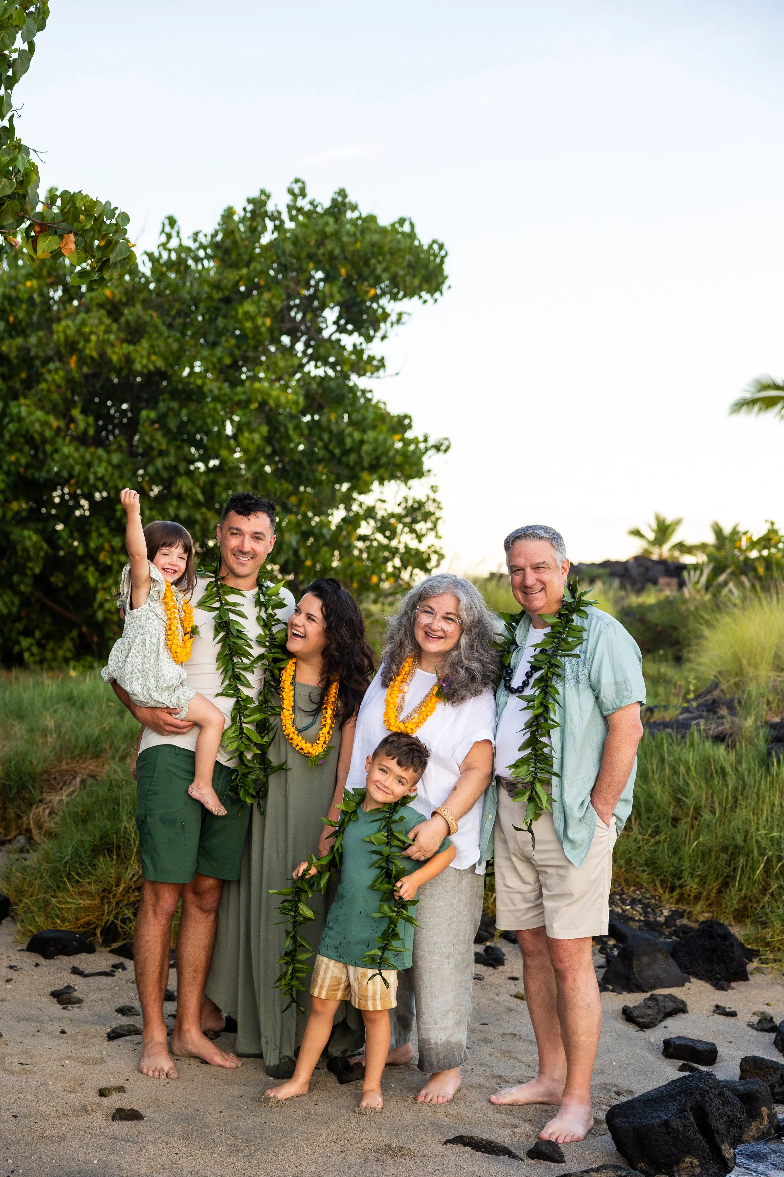 Family gathering on the beach, wearing leis, standing on sandy ground with rocks, lush greenery, and trees in the background, during sunset.