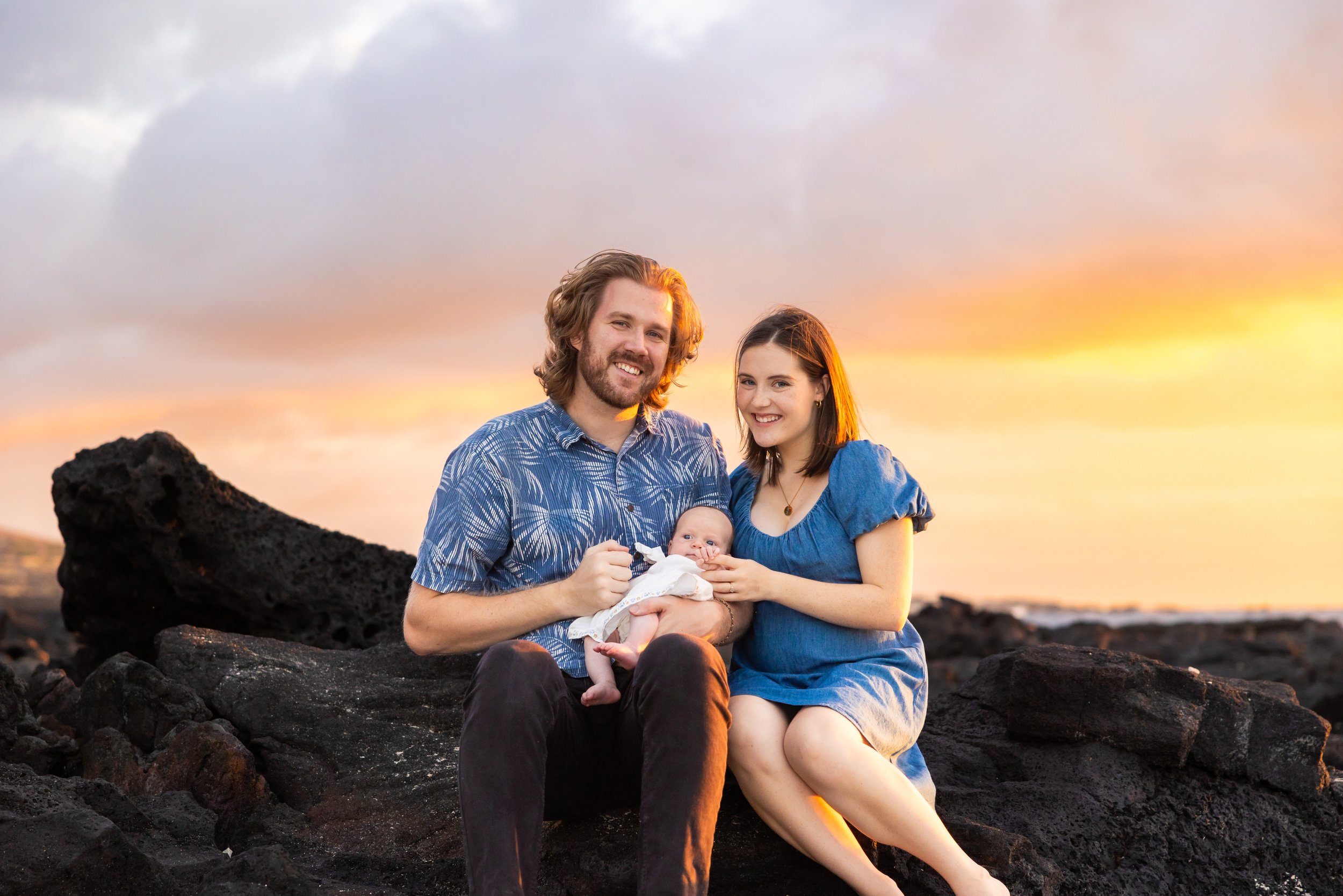 A family of three sitting on a rock together. A mom, dad, and newborn baby. The sun is setting in the background. The parents are wearing blue and the baby is wearing white.