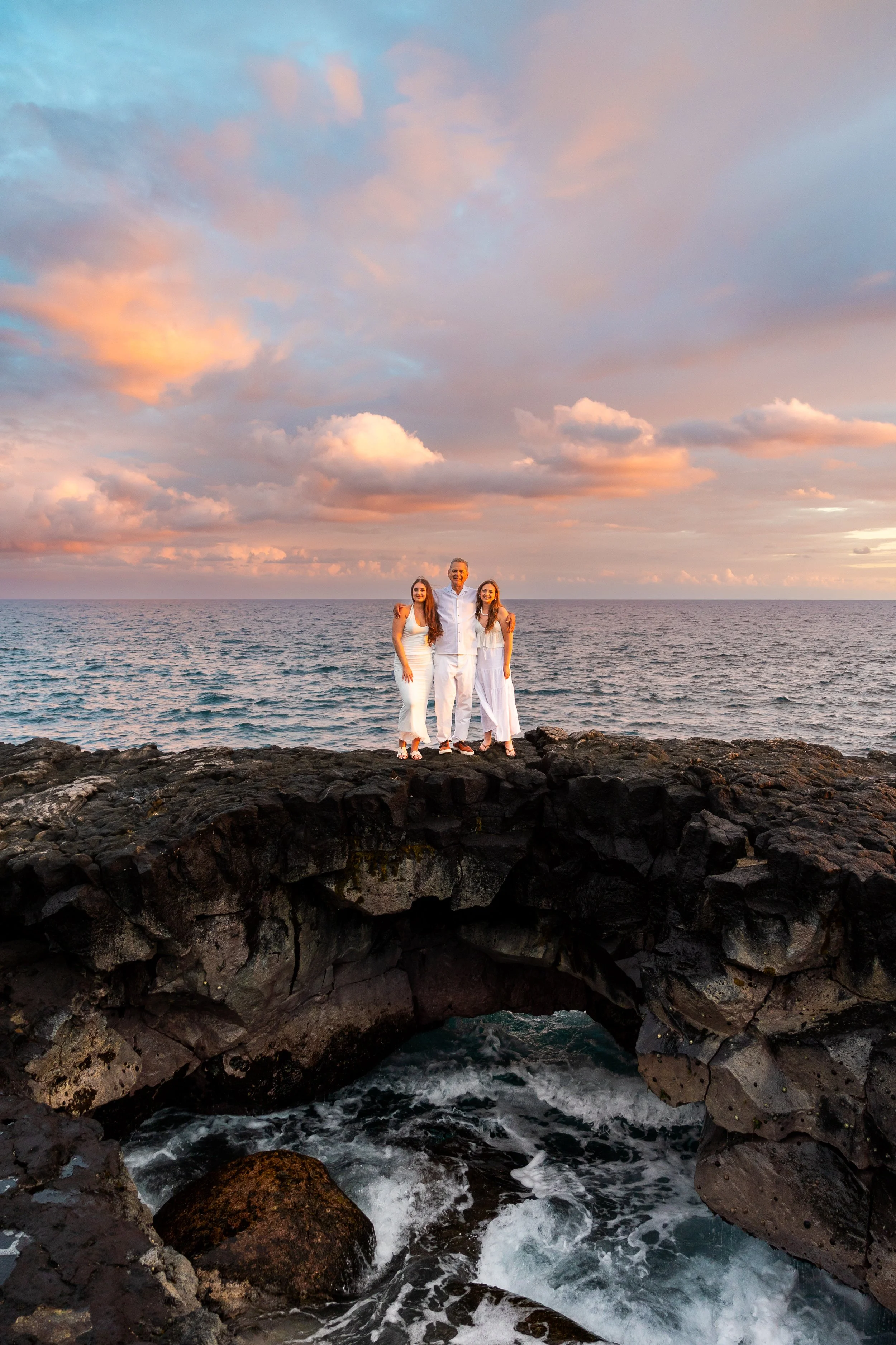 A father and his two daughters standing on a lava rock arch with the sun setting in the background. Located on the coast of Kailua-Kona.Waves from the ocean are crashing underneath the arch that the family is standing on.