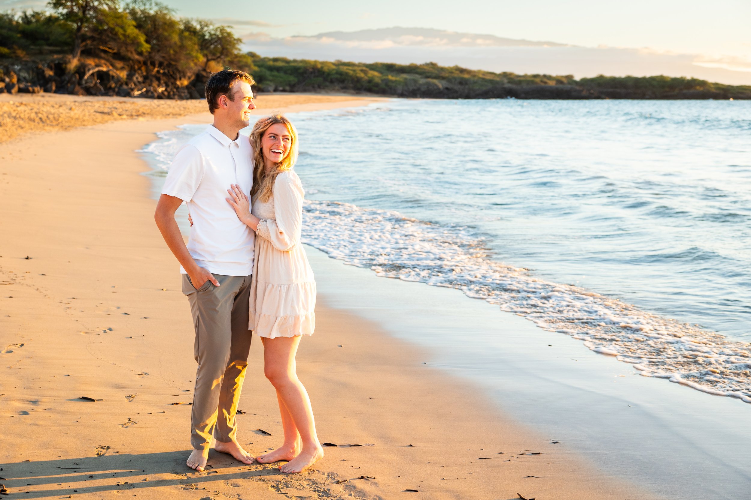 A happy couple standing on a long white sand beach. They are looking out over the ocean at sunset. Palm trees are in the background as well as the mountain Hualalai.