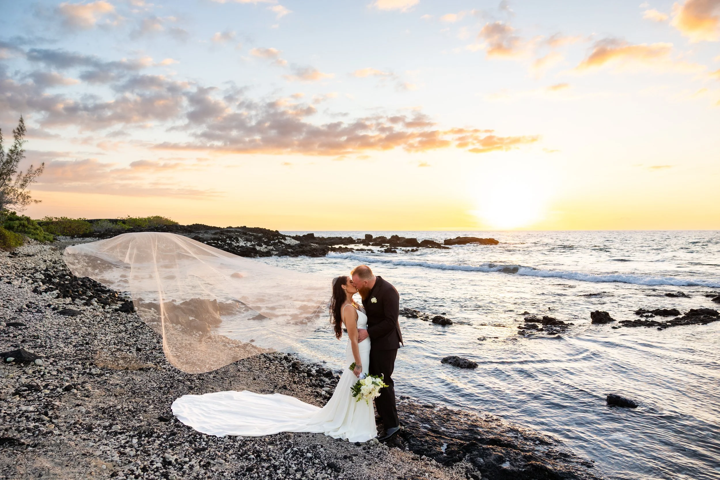 A bride and groom share a kiss on a rocky beach at sunset, with the bride wearing a white gown and holding a bouquet, and the groom in a dark suit. The bride's veil flows behind her on the ground, and the sky is filled with soft clouds and warm light