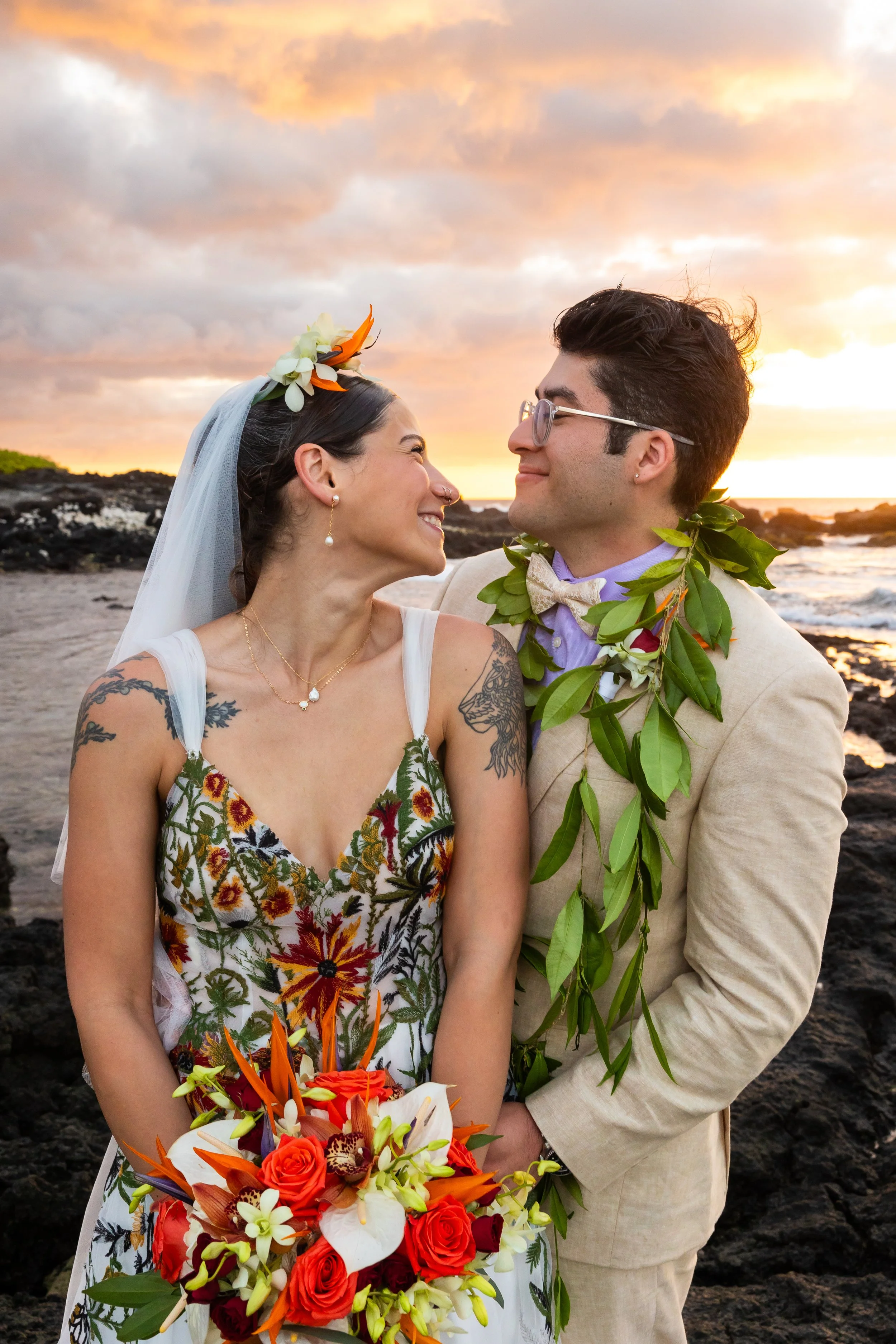 A couple dressed in wedding attire, smiling and looking into each other's eyes during sunset on a beach with clouds in the background. The bride has tattoos on her arm, wears flowers in her hair and holds a colorful bouquet. The groom wears glasses, 