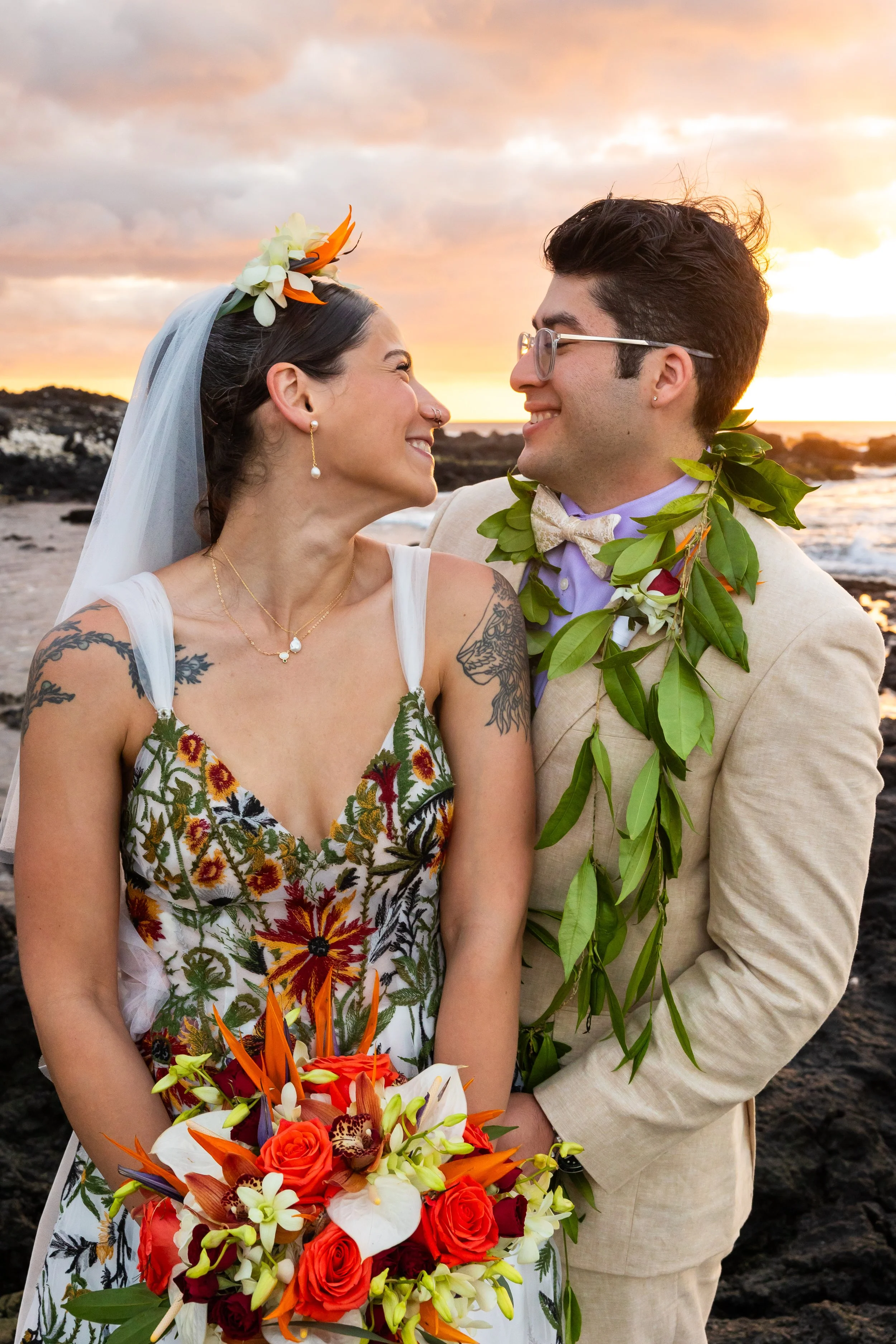 A groom in a tan suit wearing a tea leaf lei holds his bride wearing a white dress with intricate red, green and yellow details. They are on a white sand beach with lava rock on the coast of Hawaii Island. A vibrant sunset is in the background.