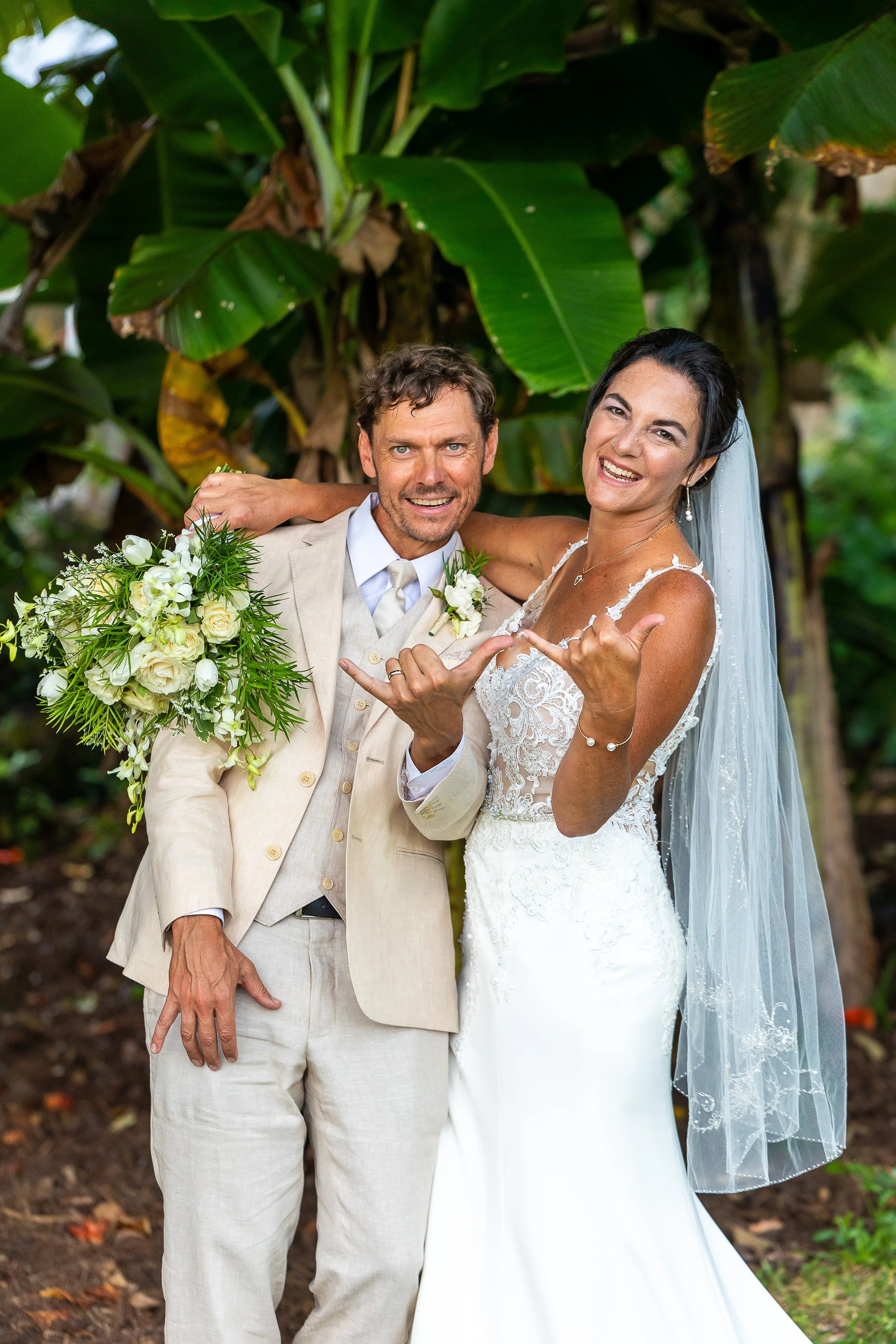 Bride and groom smiling and posing playfully in front of lush green foliage; the bride is holding a bouquet and wearing a white wedding dress and veil, while the groom is in a beige suit.