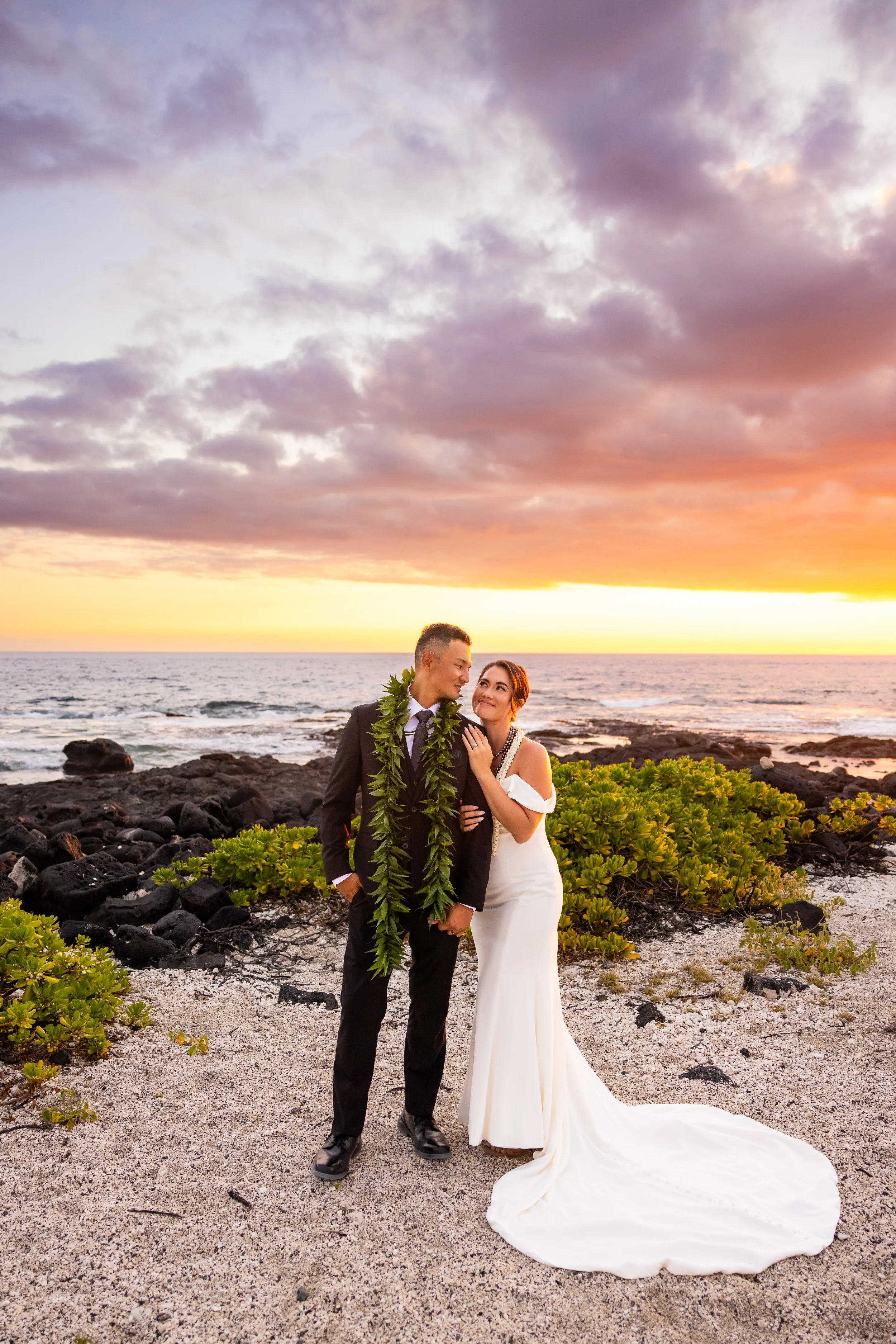 A newlywed couple stands together on a rocky beach at sunset, with the bride in a white gown and the groom in a black suit, holding hands and gazing at each other, surrounded by green plants and colorful clouds in the sky.