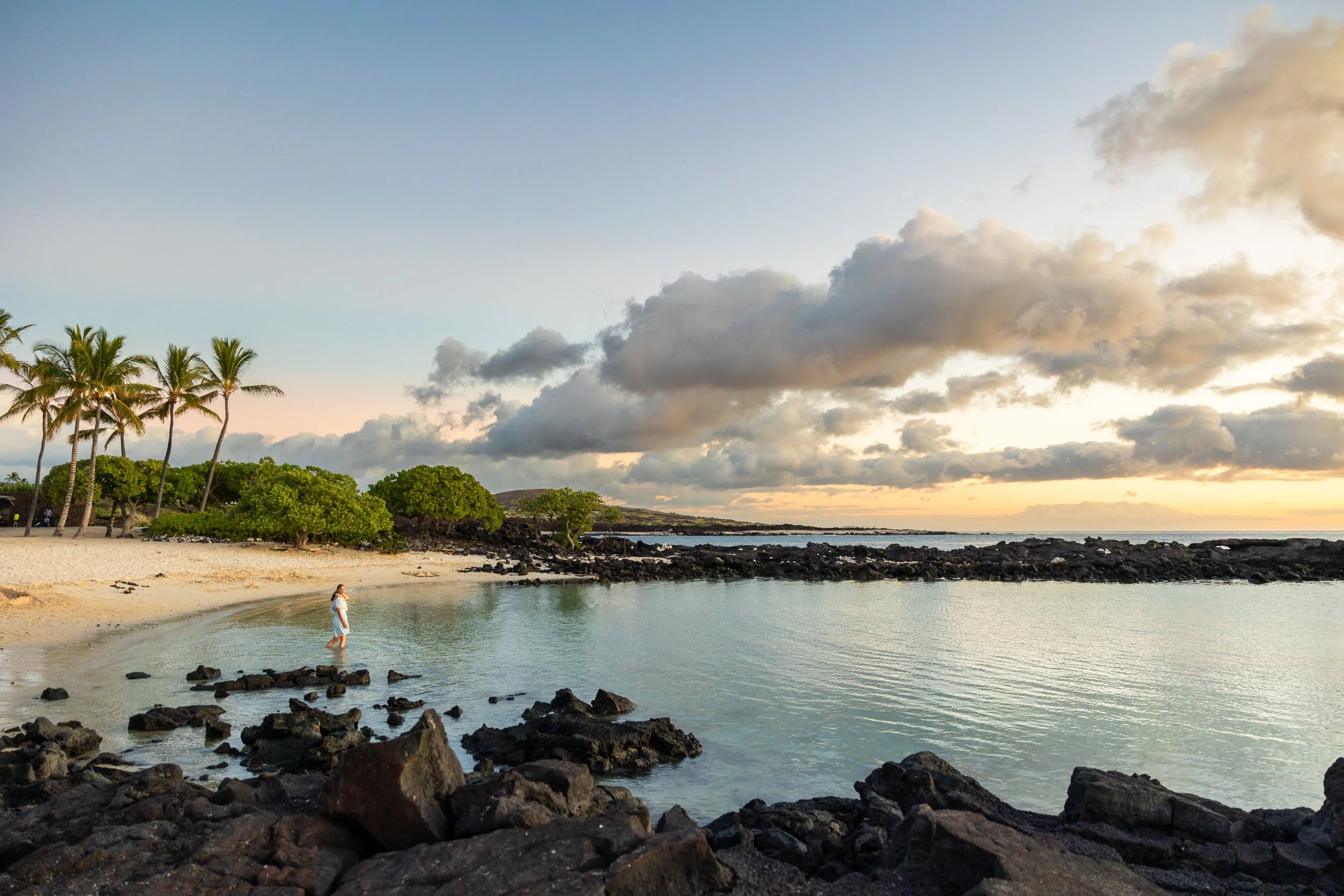 White sandy beach at sunset with lava rocks inthe foreground and background. A girl is posing for a photo with palm trees in the background. Located near Waikoloa Hawaii