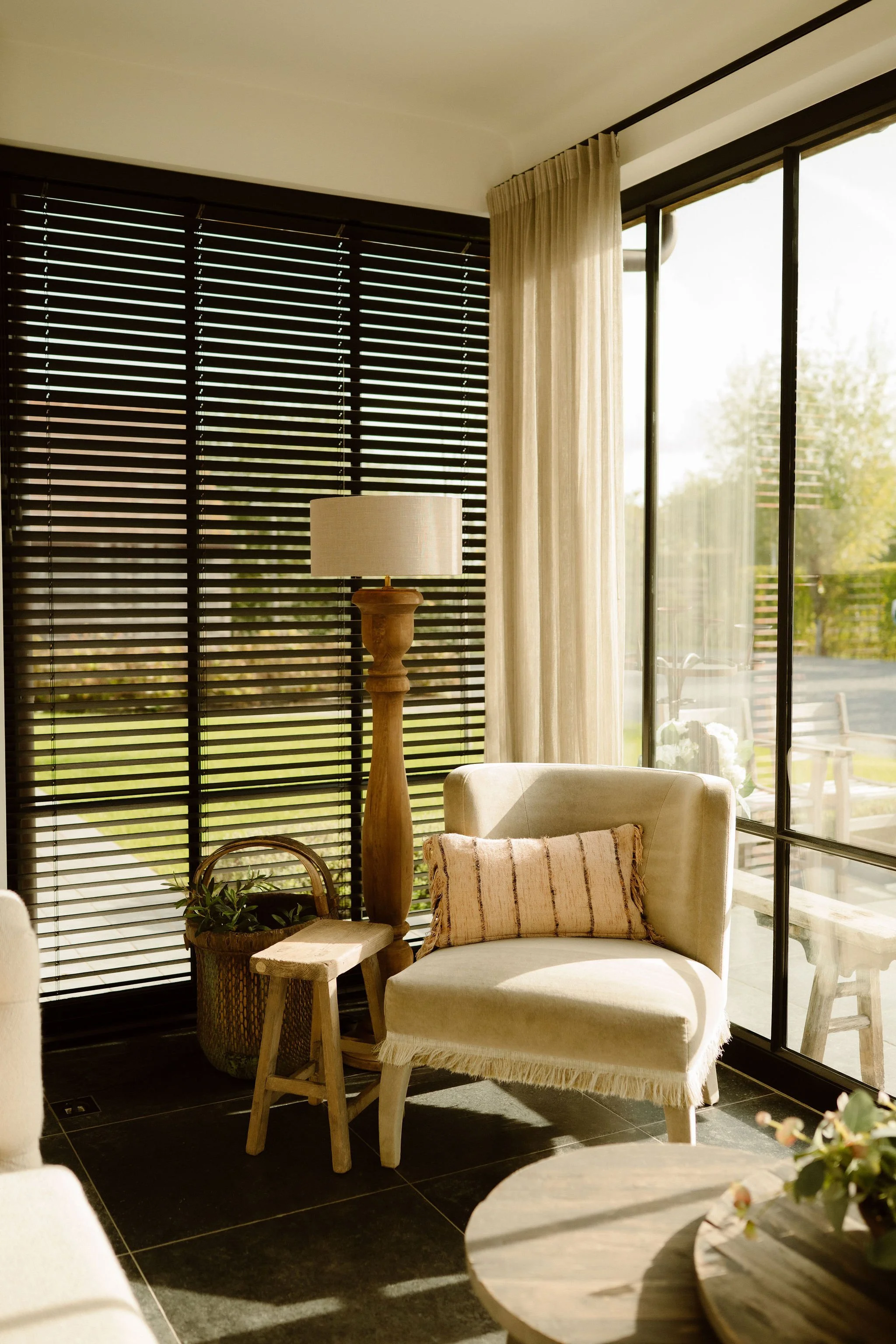 Een woonkamer met grote ramen en houten jaloezieën, een beige stoel met een gestreepte kussen, een houten tafel en een vloerlamp, veel natuurlijk licht en een tuin buiten zichtbaar.
