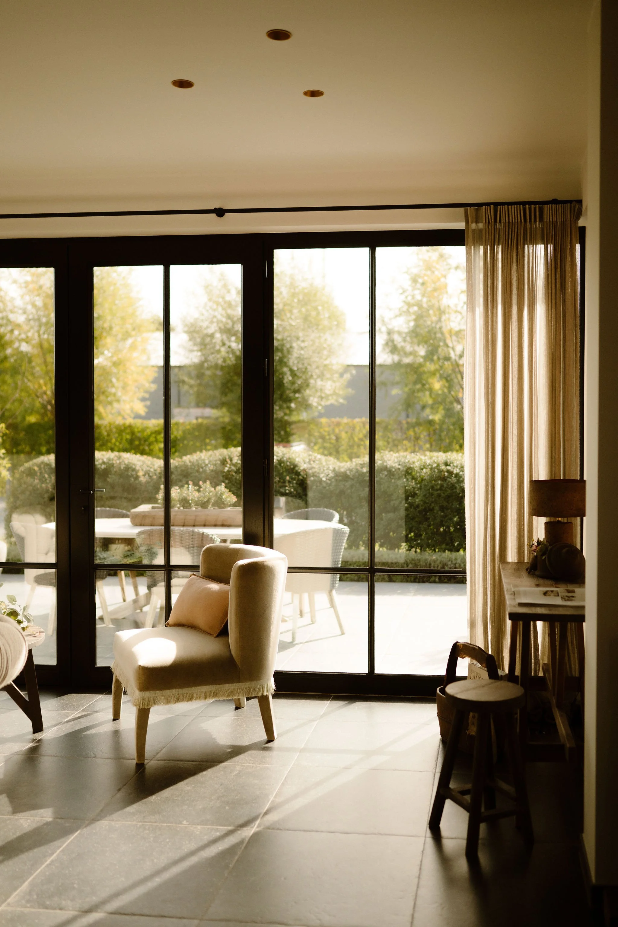 Orangerie met uitzicht op de tuin - Het Verborgen Geluk, adEen gezellige woonkamer met grote ramen en curtains, een beige bank, een blauwe fauteuil met een beige kussen, een ronde houten salontafel met decoraties, en een grote plant in een zwart pot.