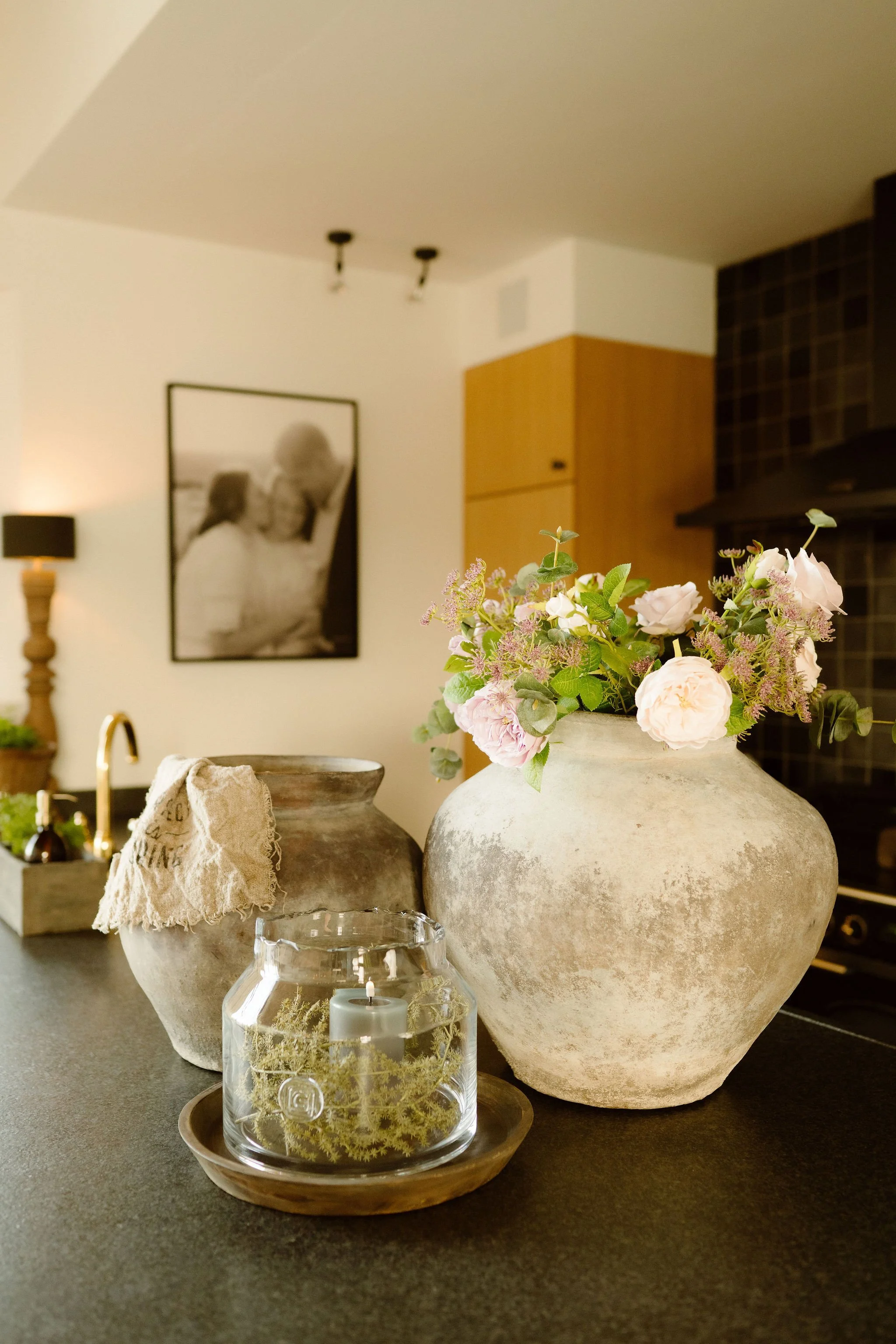 Een kamer met een bloemenvaas, een kaars in een glazen houder, een ander vaas en een doek op een tafel. Op de achtergrond hangt een foto, en rechts een keukenmuur en kast.