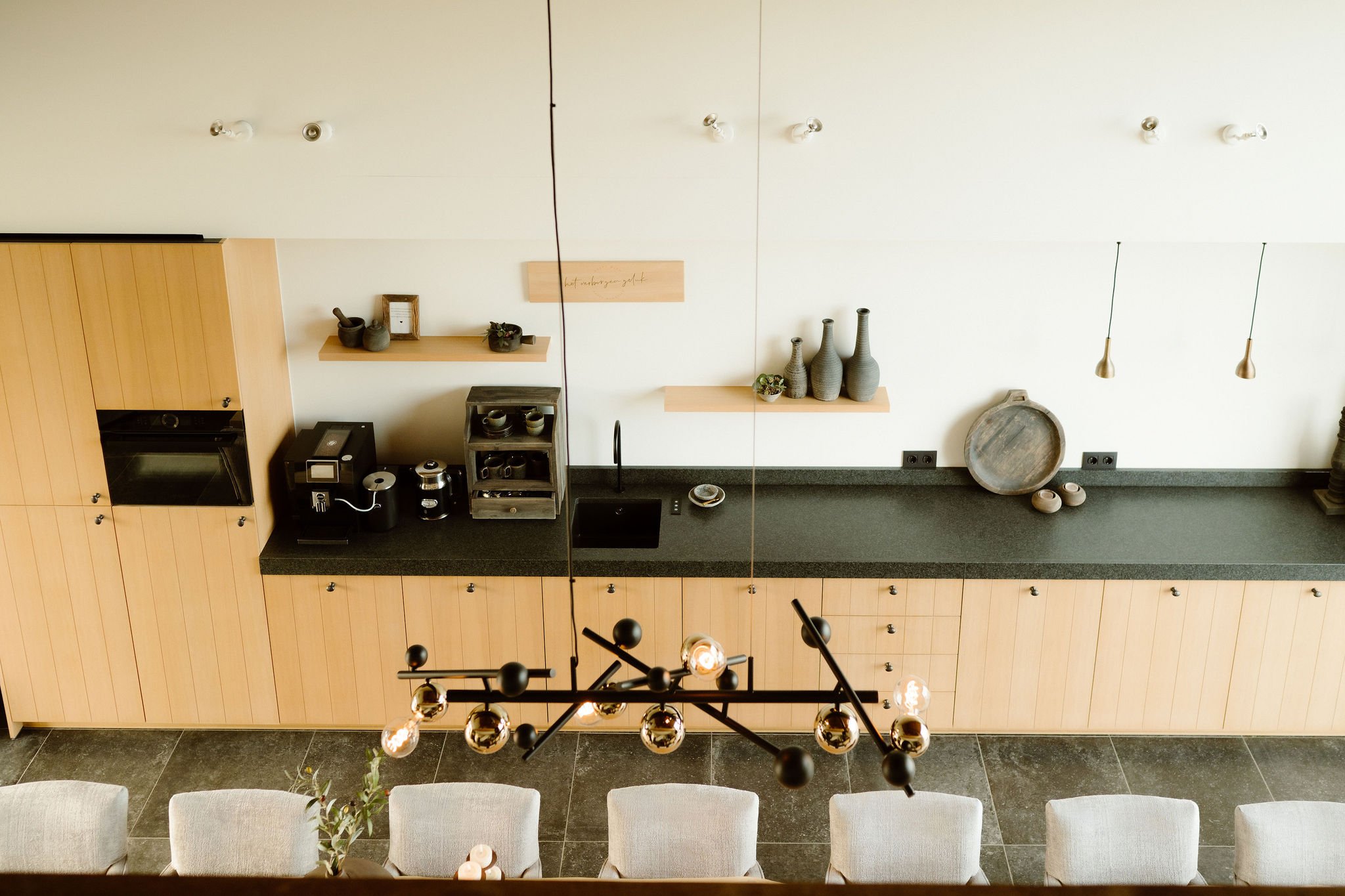 Een keuken met houten kasten, een black granite werkblad, en decoratieve vazen en potten op de muur. Er hangt een decoratieve lamp aan het plafond. De vloer is van donkersteeen.