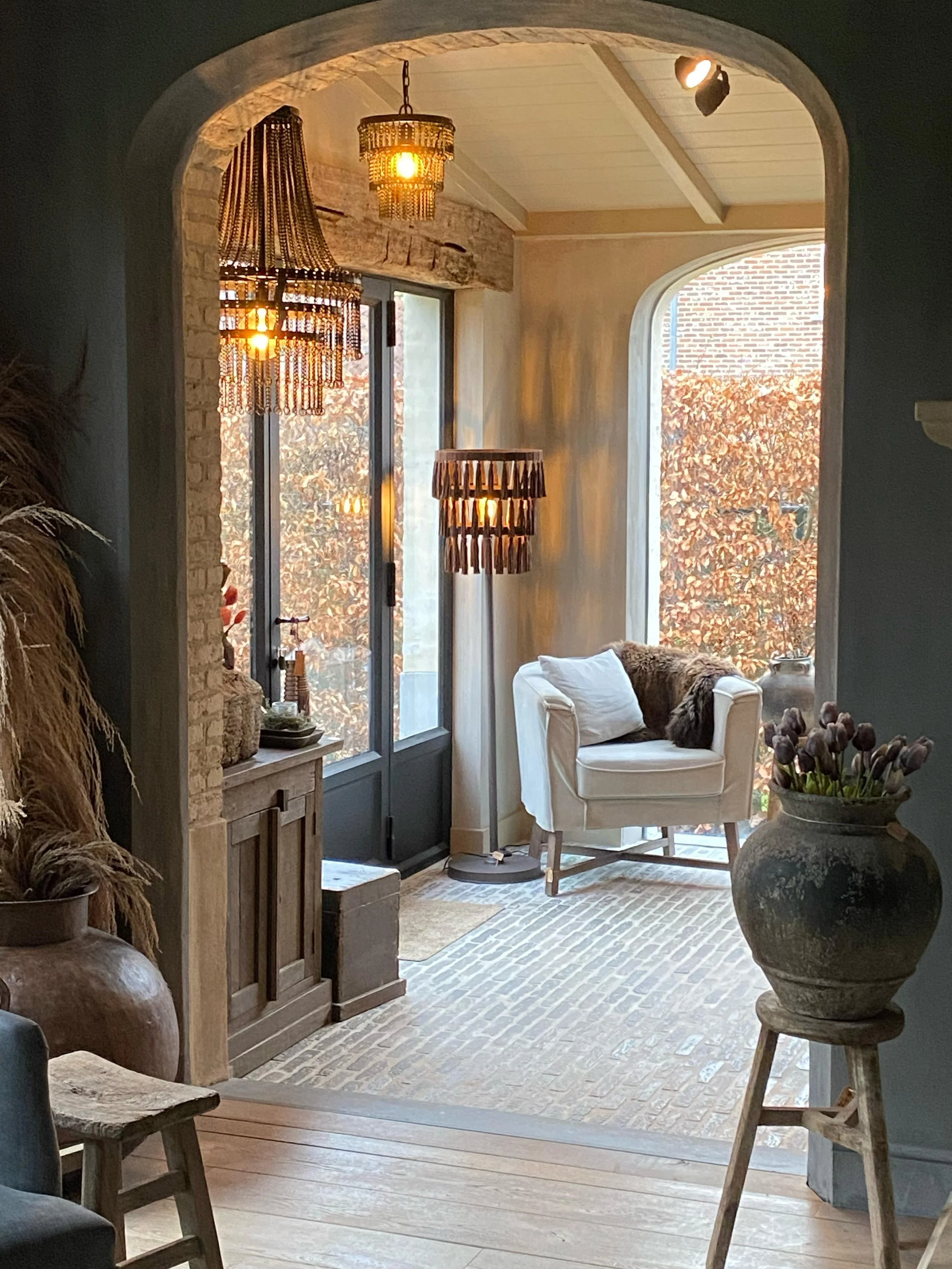 Gezellige zithoek met witte fauteuil, sierkussens en zachte verlichting, gelegen bij grote ramen met uitzicht op herfstbladeren.