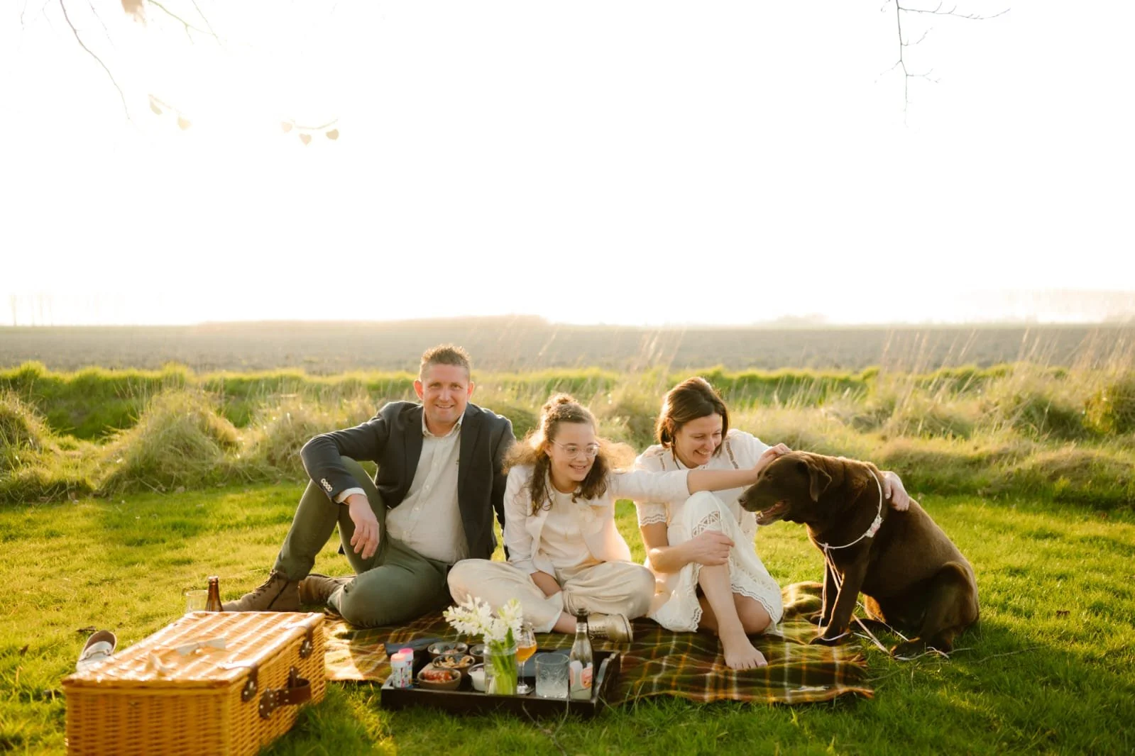 Familie geniet van een picknick op een grasveld tijdens zonsondergang met een hond en verzorgingsproducten op een picknickkleed.