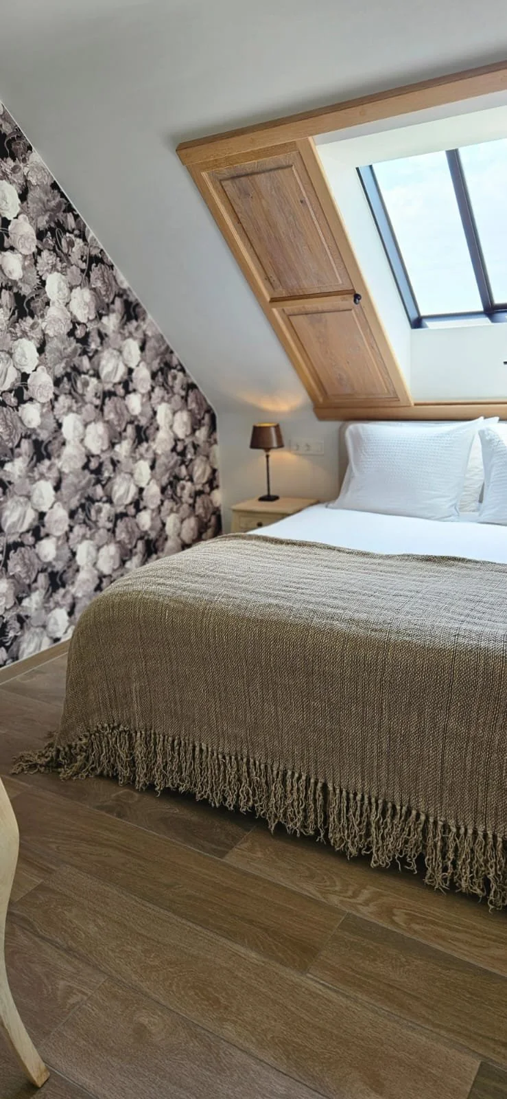 Slaapkamer met wit beddengoed, beige bedsprei, een zijtafel met lamp, een houten plafondluik en een groot dakraam voor natuurlijk licht.