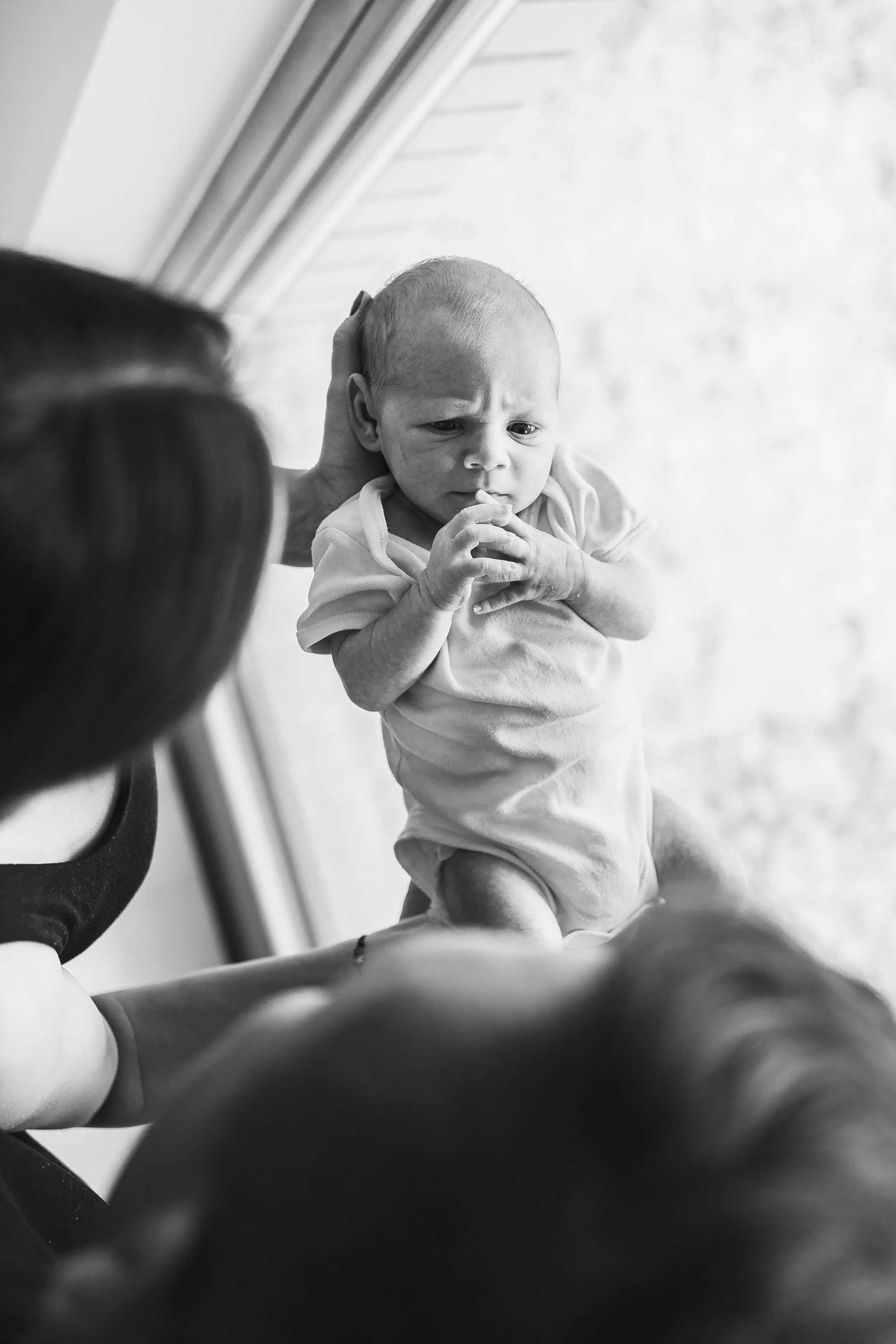 A baby holding their hands together and looking worried, being supported by an adult's hands, with another person partially visible in the foreground, near a window.