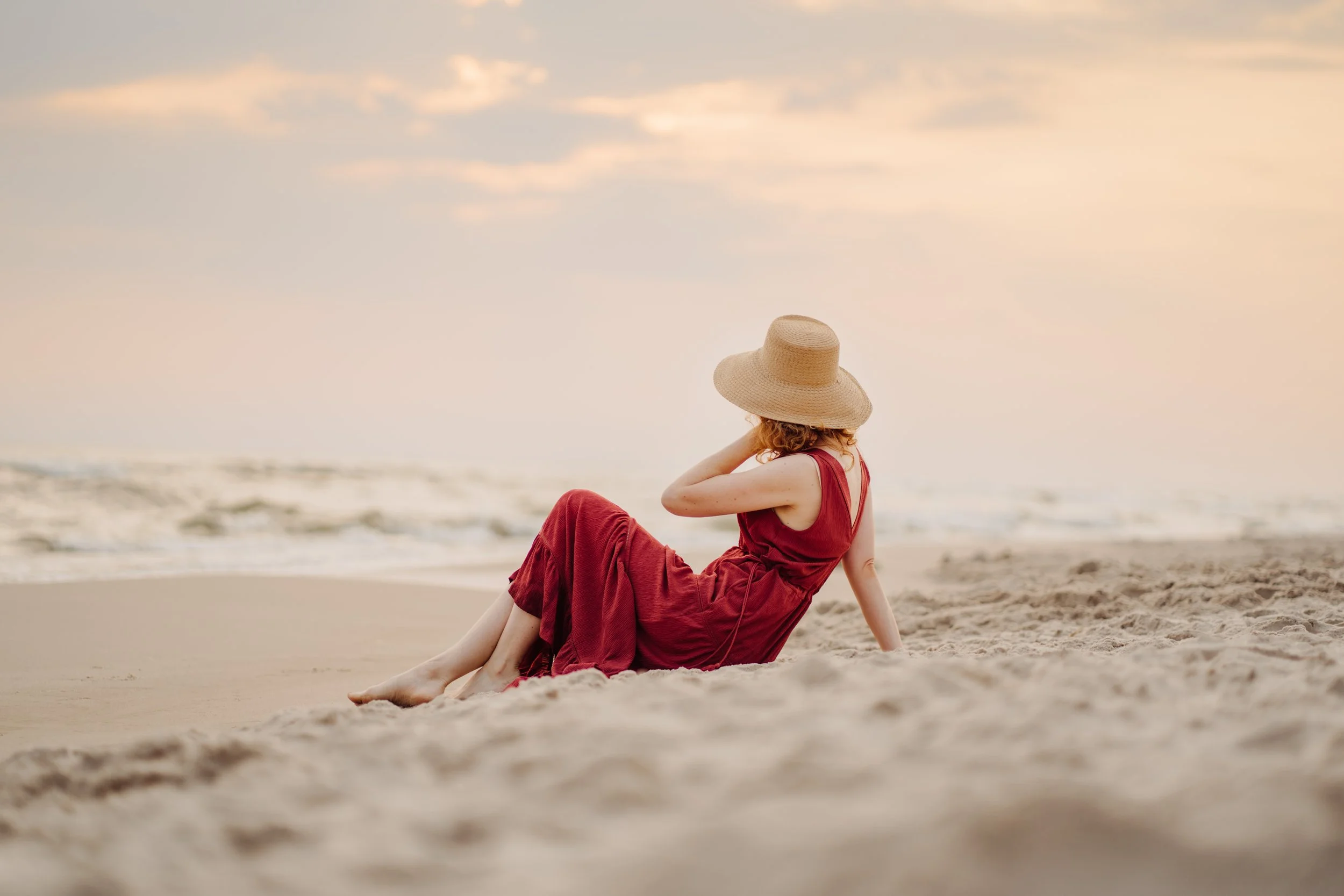 Woman in a red dress sitting on the beach, wearing a wide-brimmed straw hat, looking at the ocean during sunset.