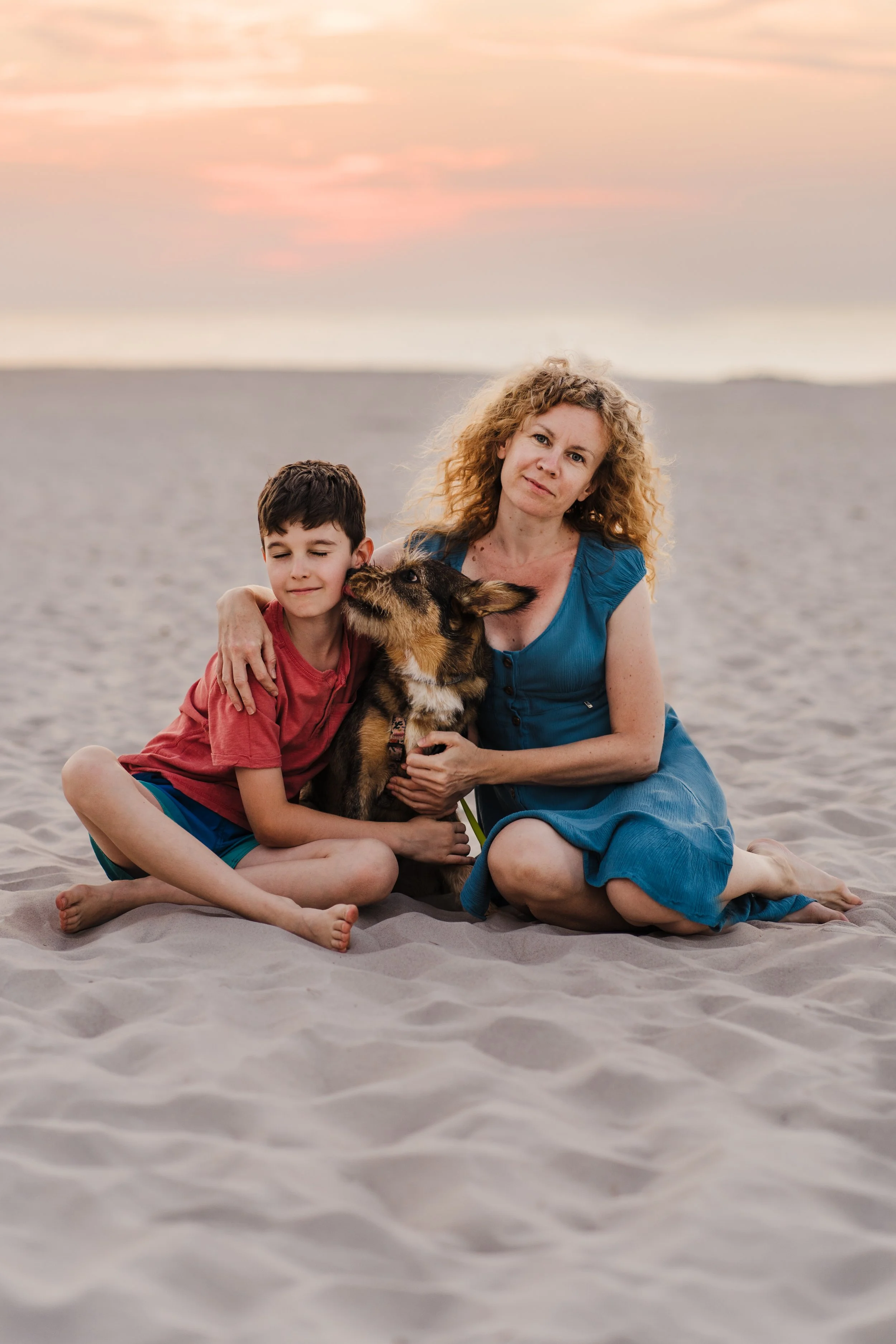 A woman, a boy, and a dog sitting on a sandy beach at sunset, with the woman and boy hugging and the dog being licked by the boy.