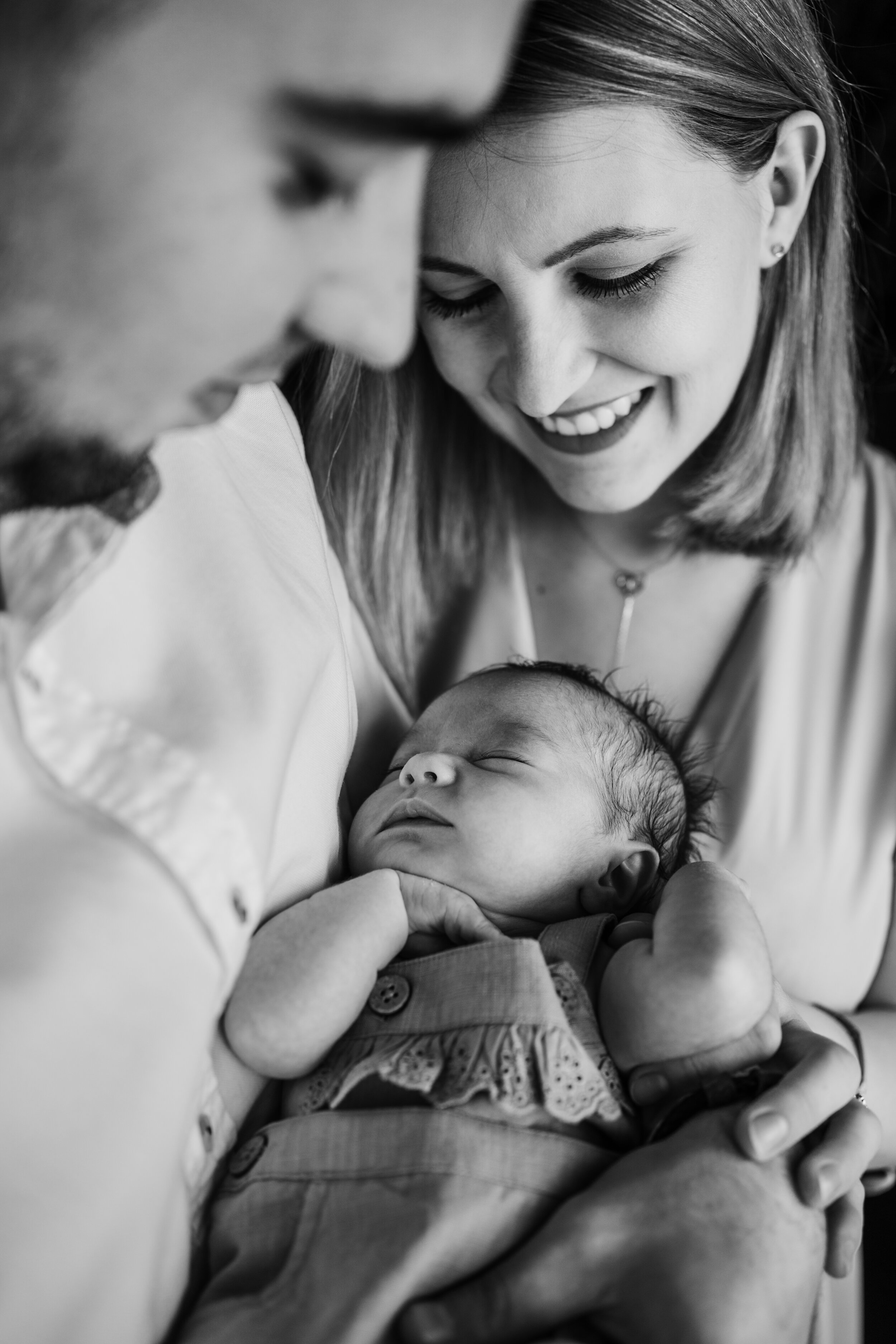 A black-and-white photo of a woman and a man holding a sleeping baby, smiling as they look down at the baby.